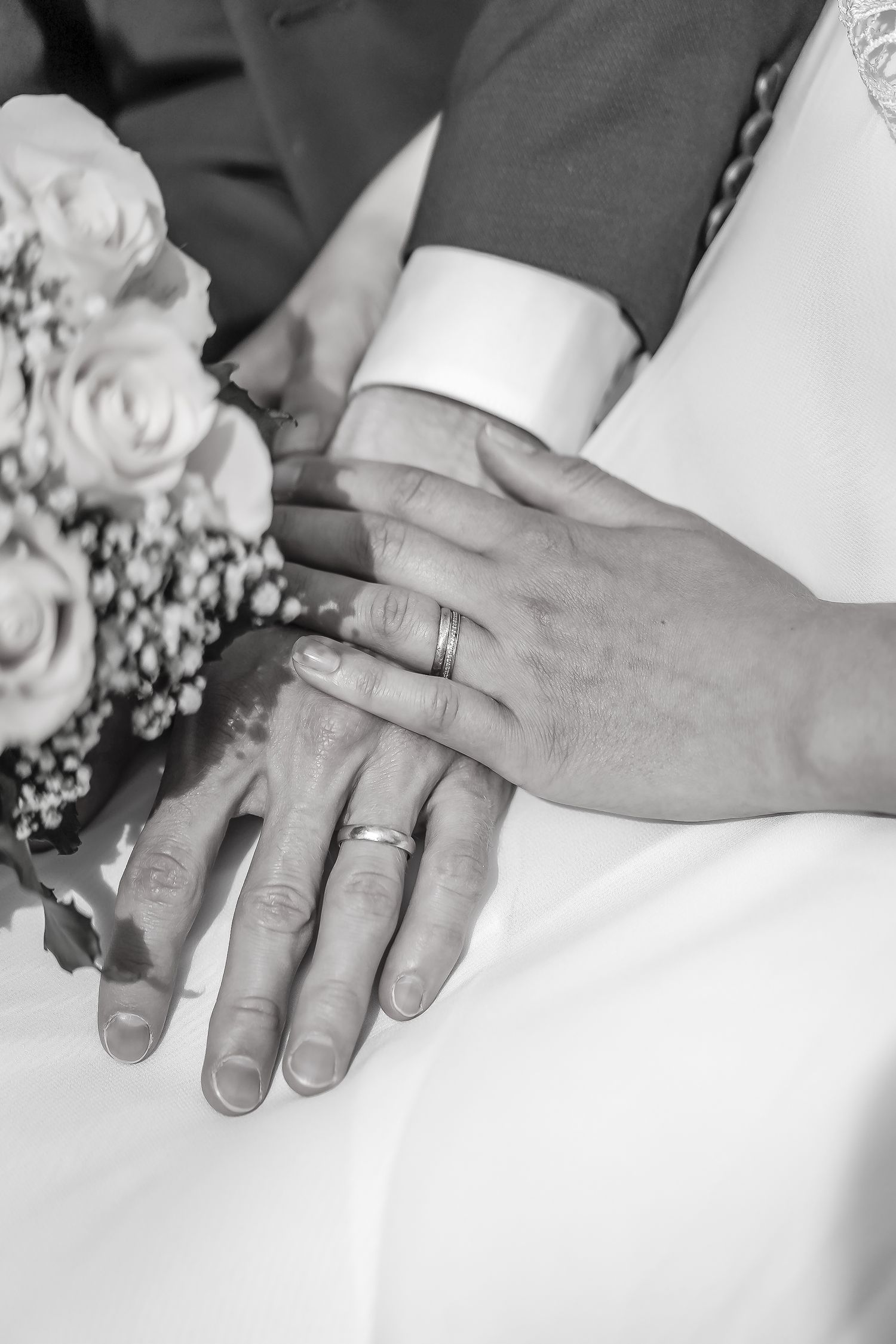 Black and white photo of newlywed couple showing wedding rings on hands with bridal bouquet nearby.