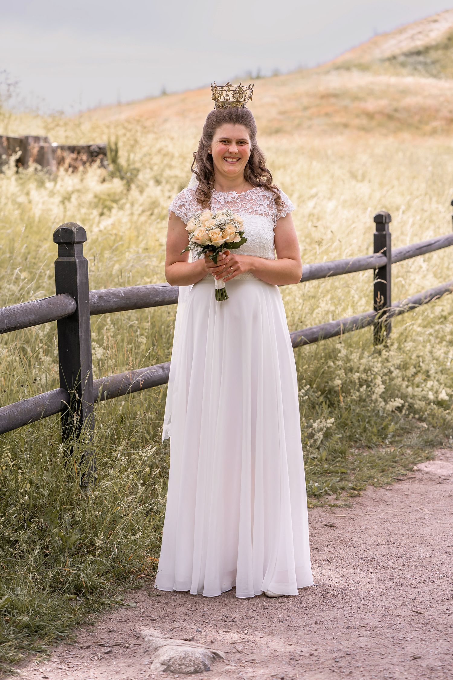 Bride in white gown and crown holds bouquet by rustic fence in field.