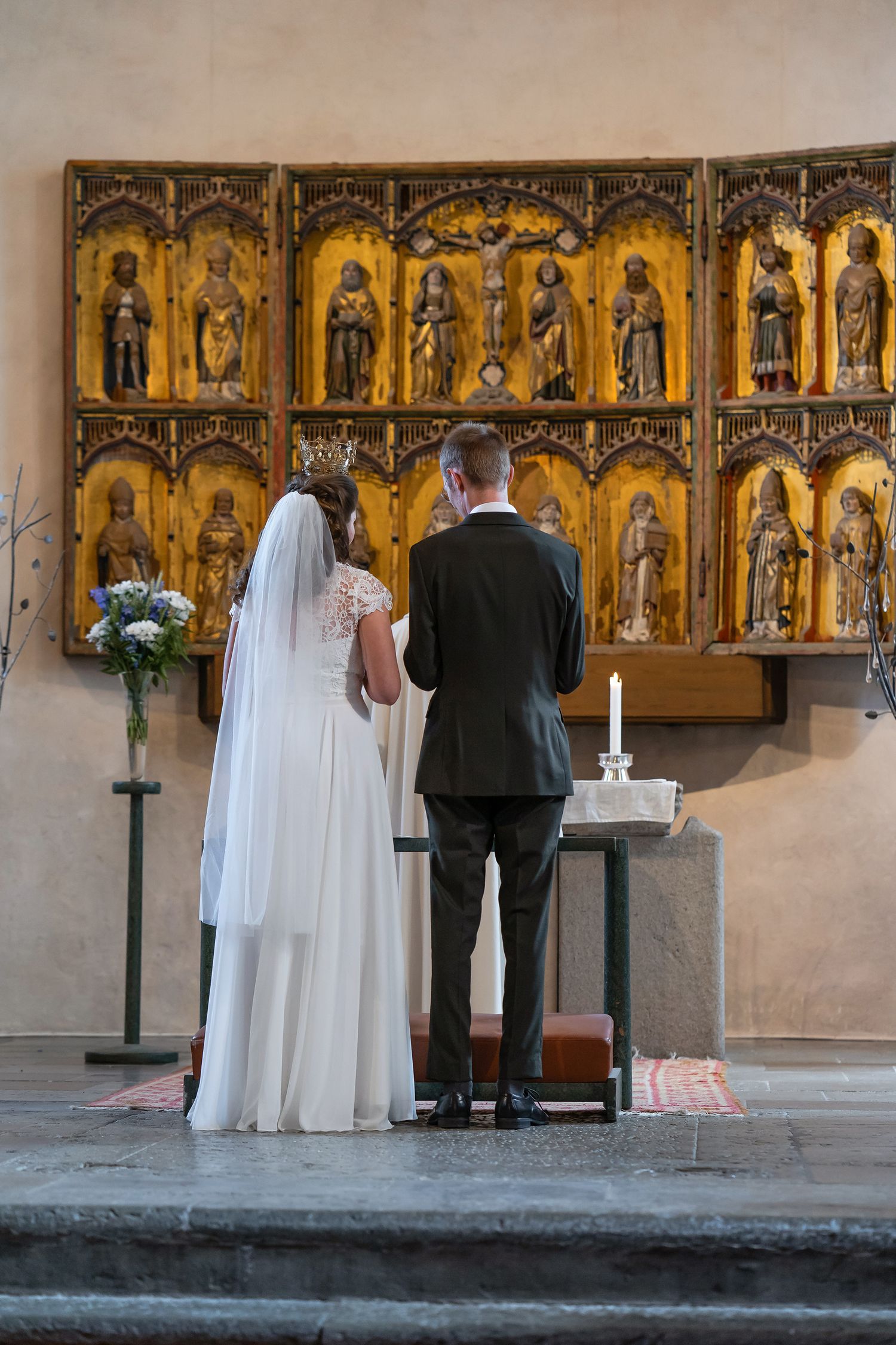 Bride and groom standing at church altar before ornate golden altarpiece during wedding ceremony.