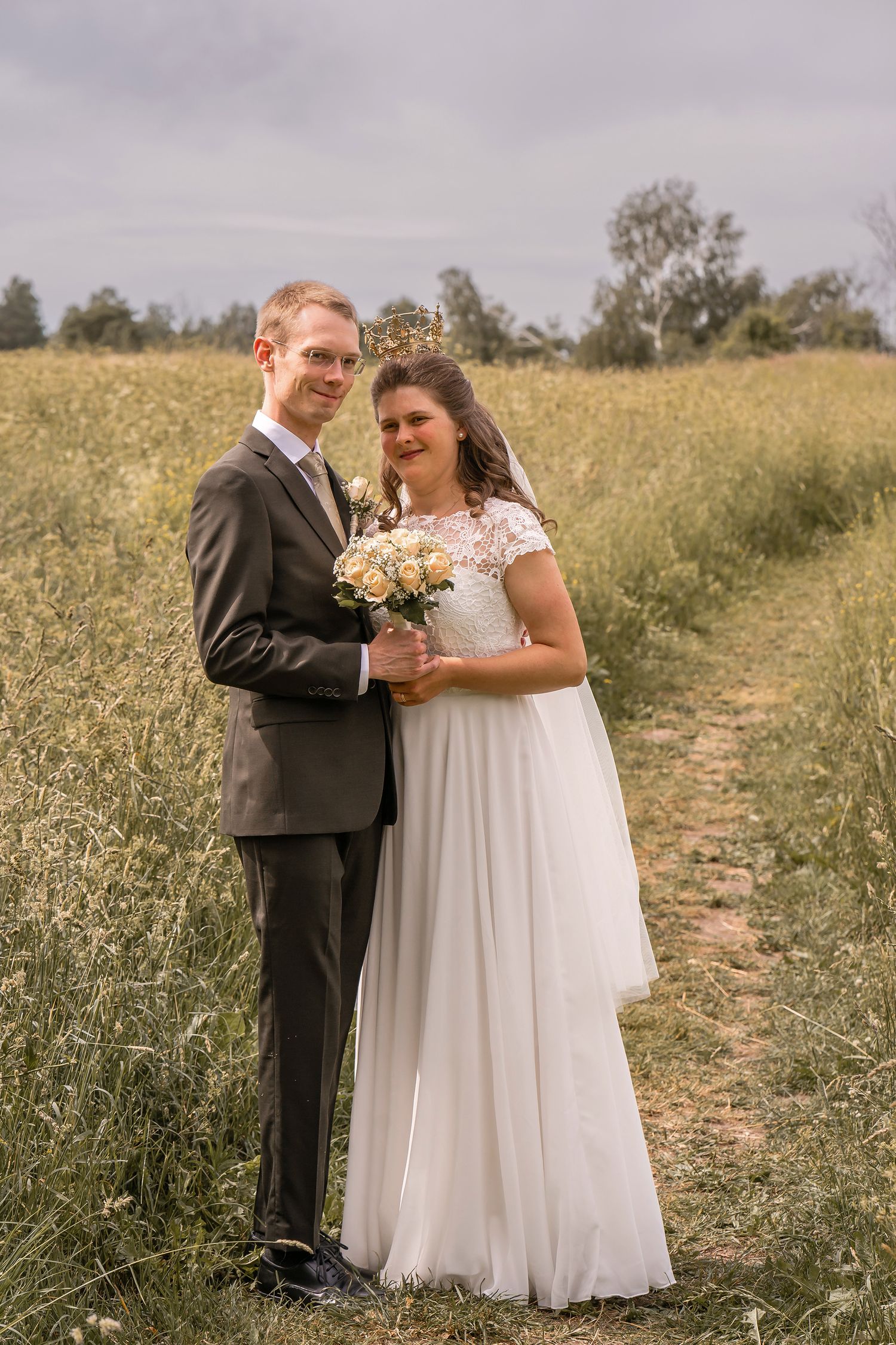 A bride and groom pose together in a golden field, she holds a white bouquet wearing a lace gown, he wears a gray suit.