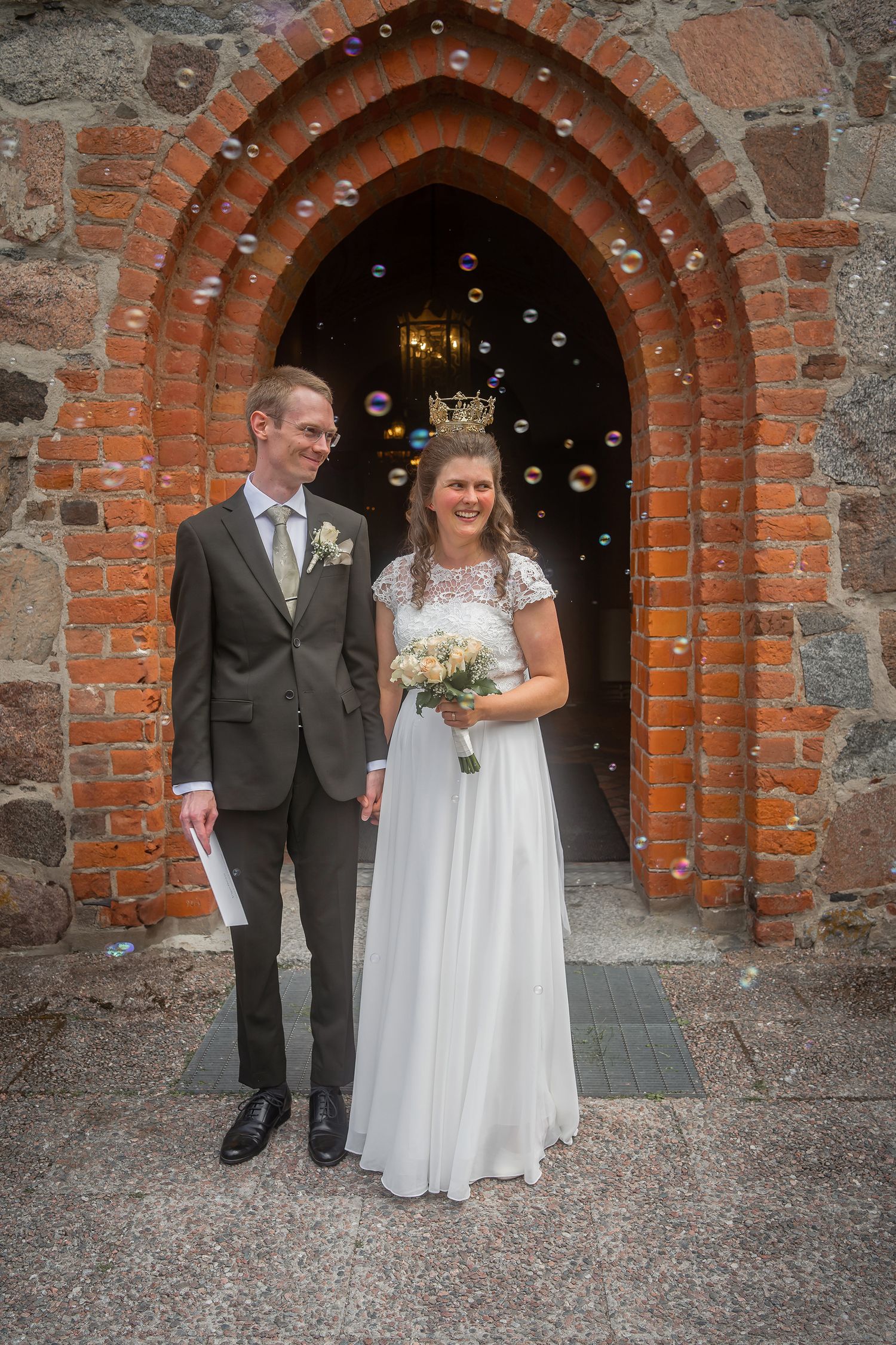 Bride and groom posing in brick church archway with soap bubbles, bride holding bouquet in white gown, groom in gray suit.