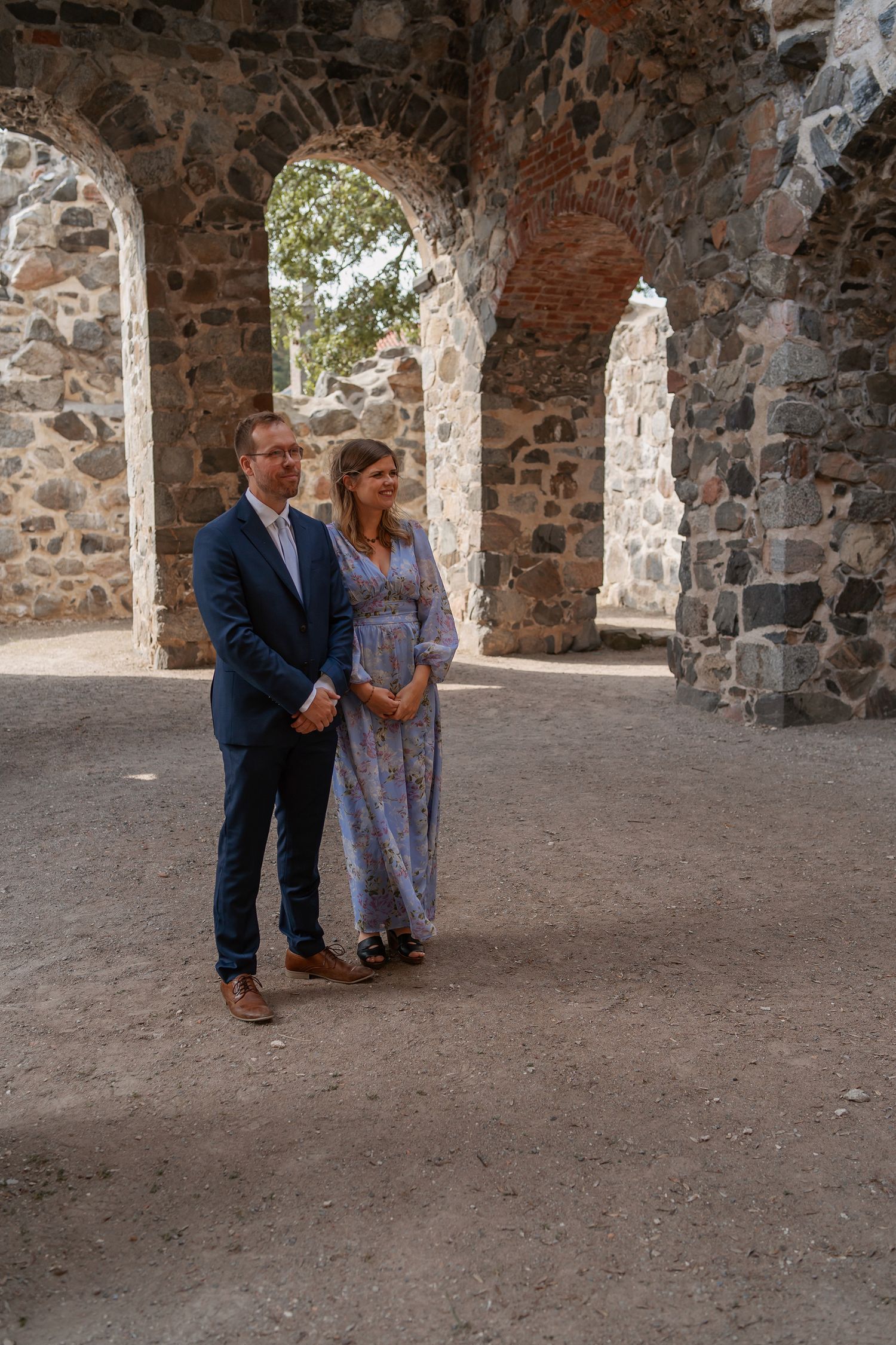 Couple in formal attire stands together inside historic stone ruins with arched doorways during wedding event.
