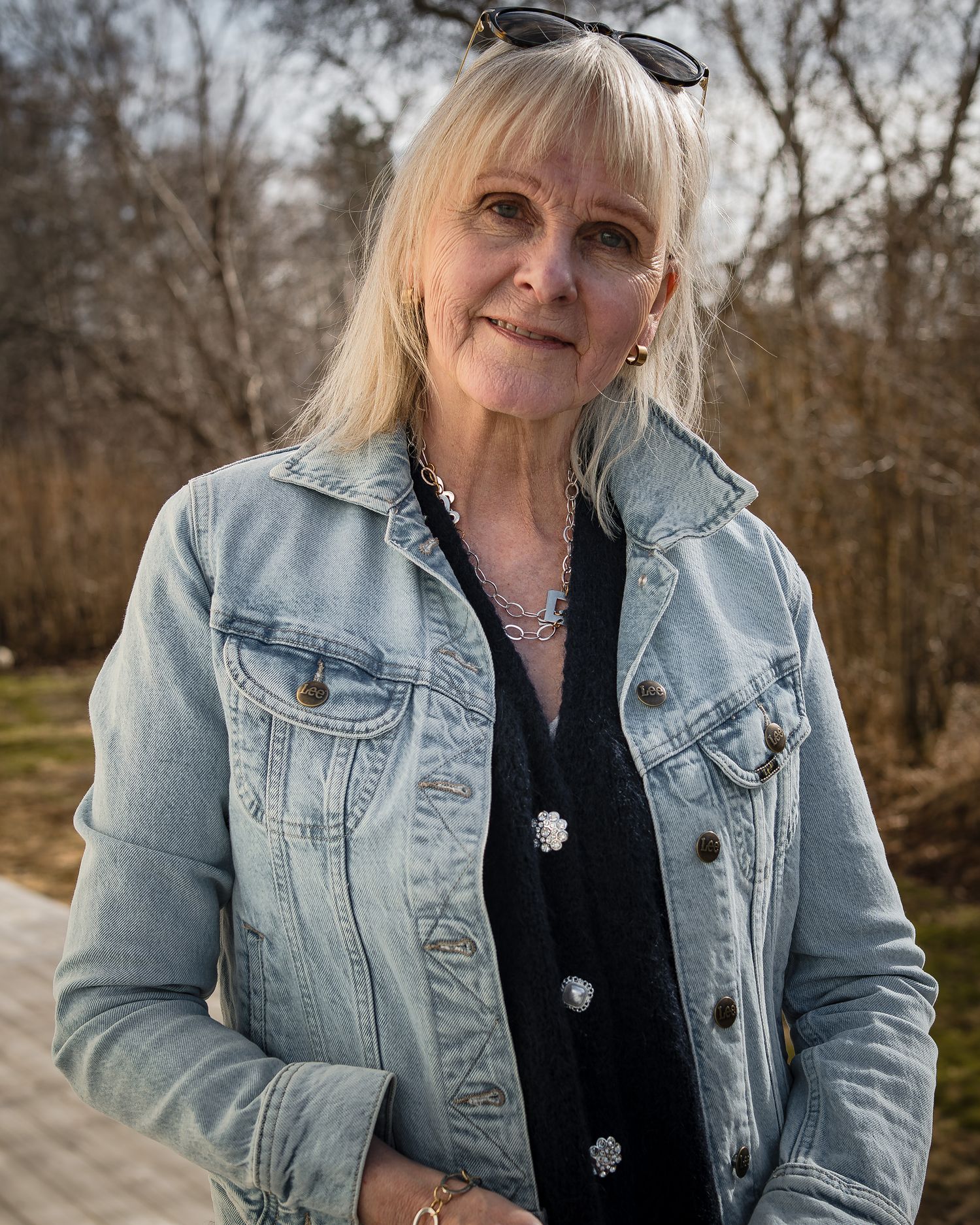 A person wearing a light denim jacket and black top poses outdoors against a natural wooded background.