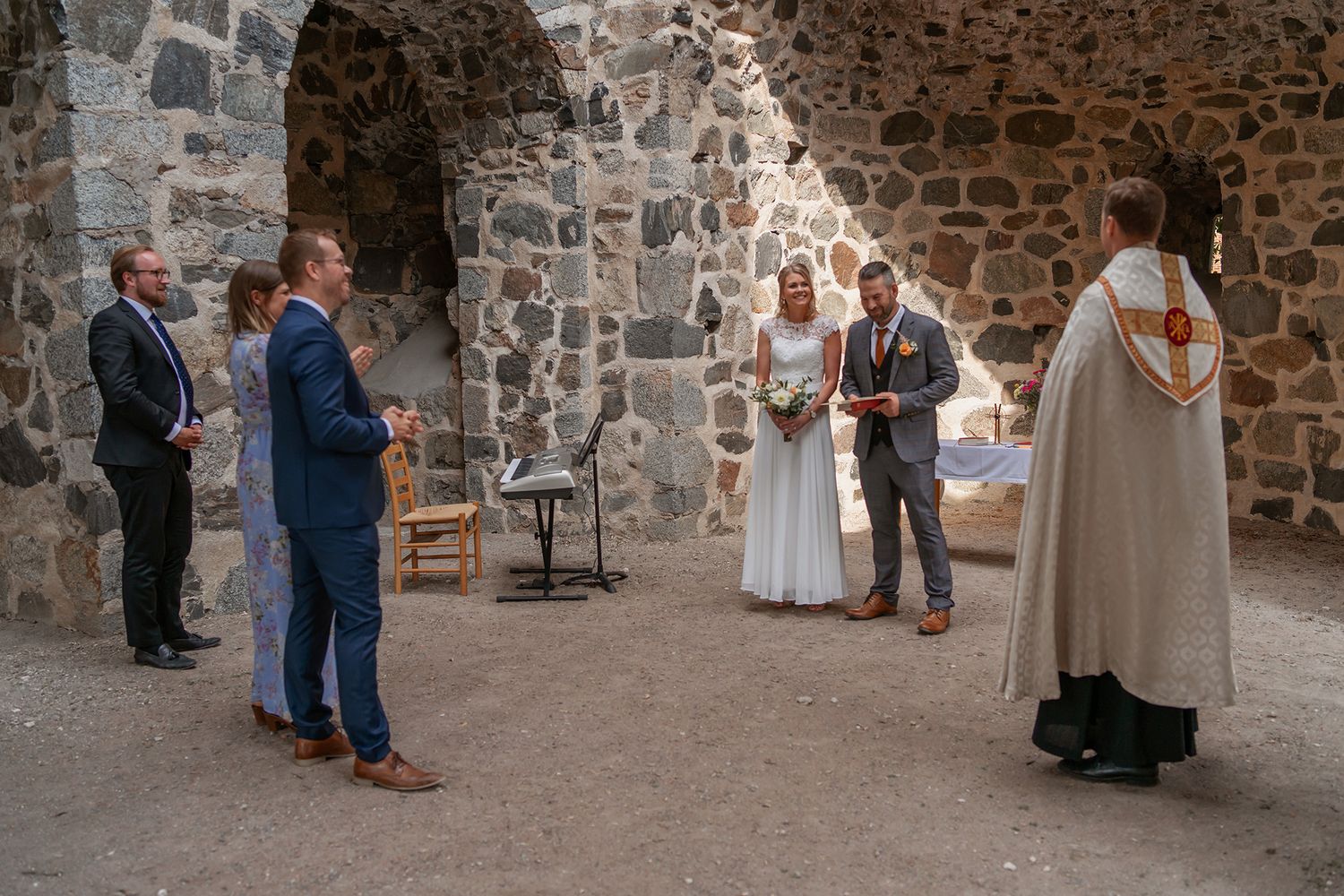 Wedding ceremony in ancient stone ruins with bride, groom, officiant, and two guests standing in sunlit space.