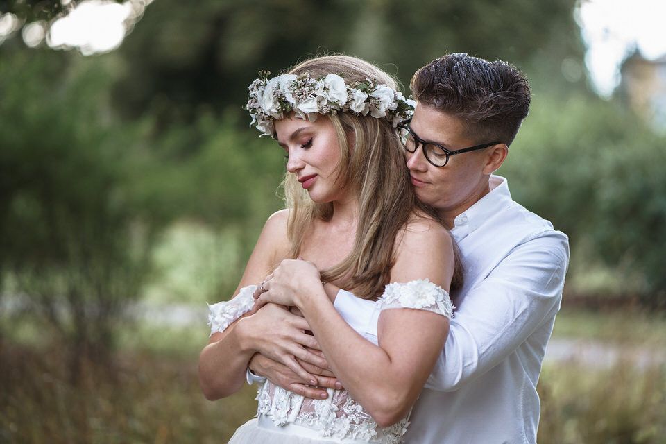 A couple in wedding attire embraces outdoors, the bride wearing a floral crown and lace dress, groom in white shirt with glasses.