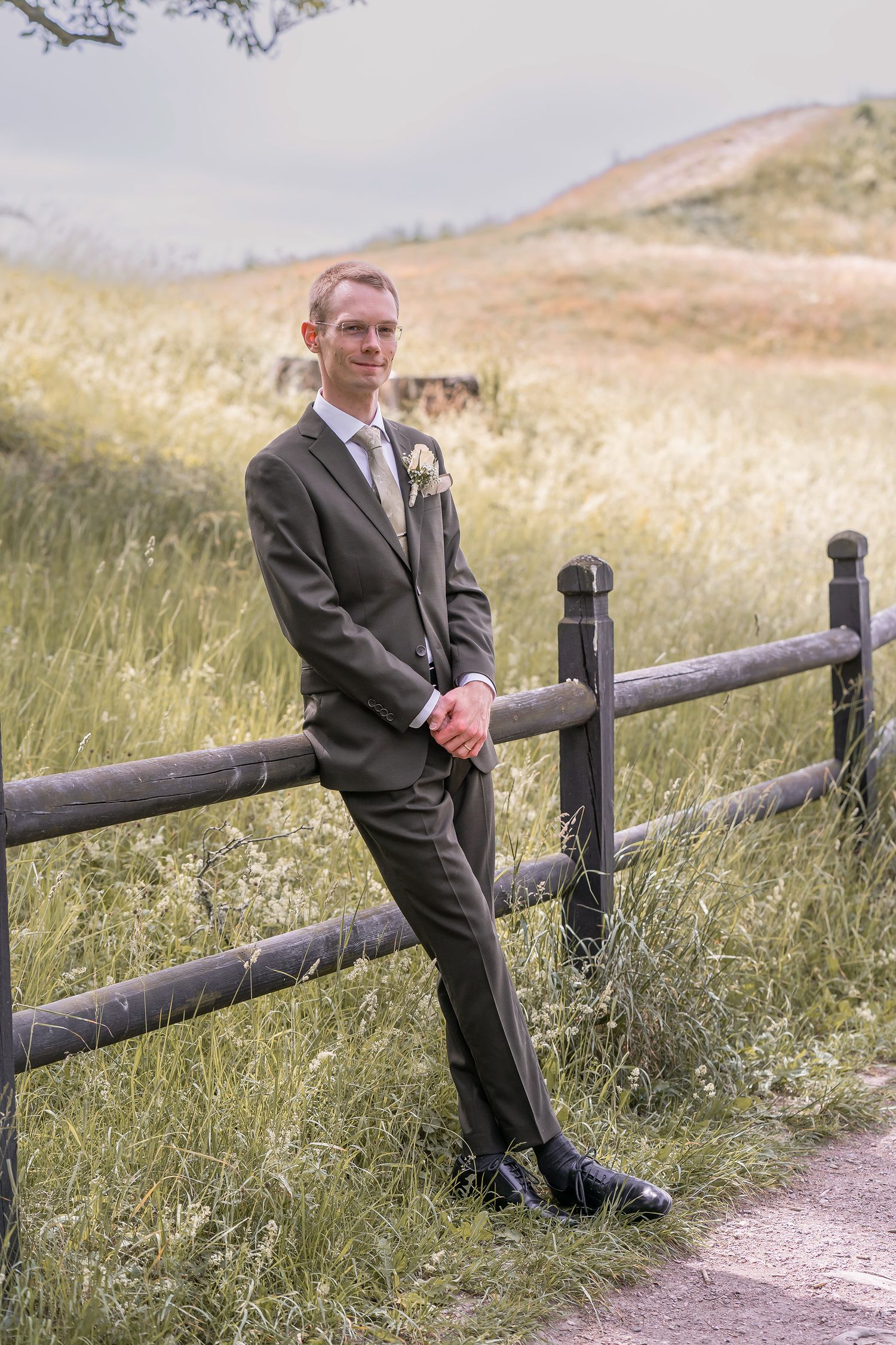 Groom in gray suit with boutonniere leaning against wooden fence in scenic countryside field on wedding day.