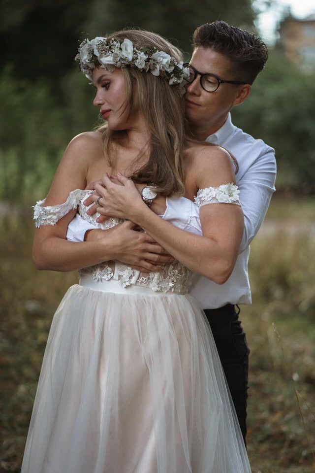 Groom embraces bride in white floral wedding dress and flower crown in romantic outdoor garden setting.