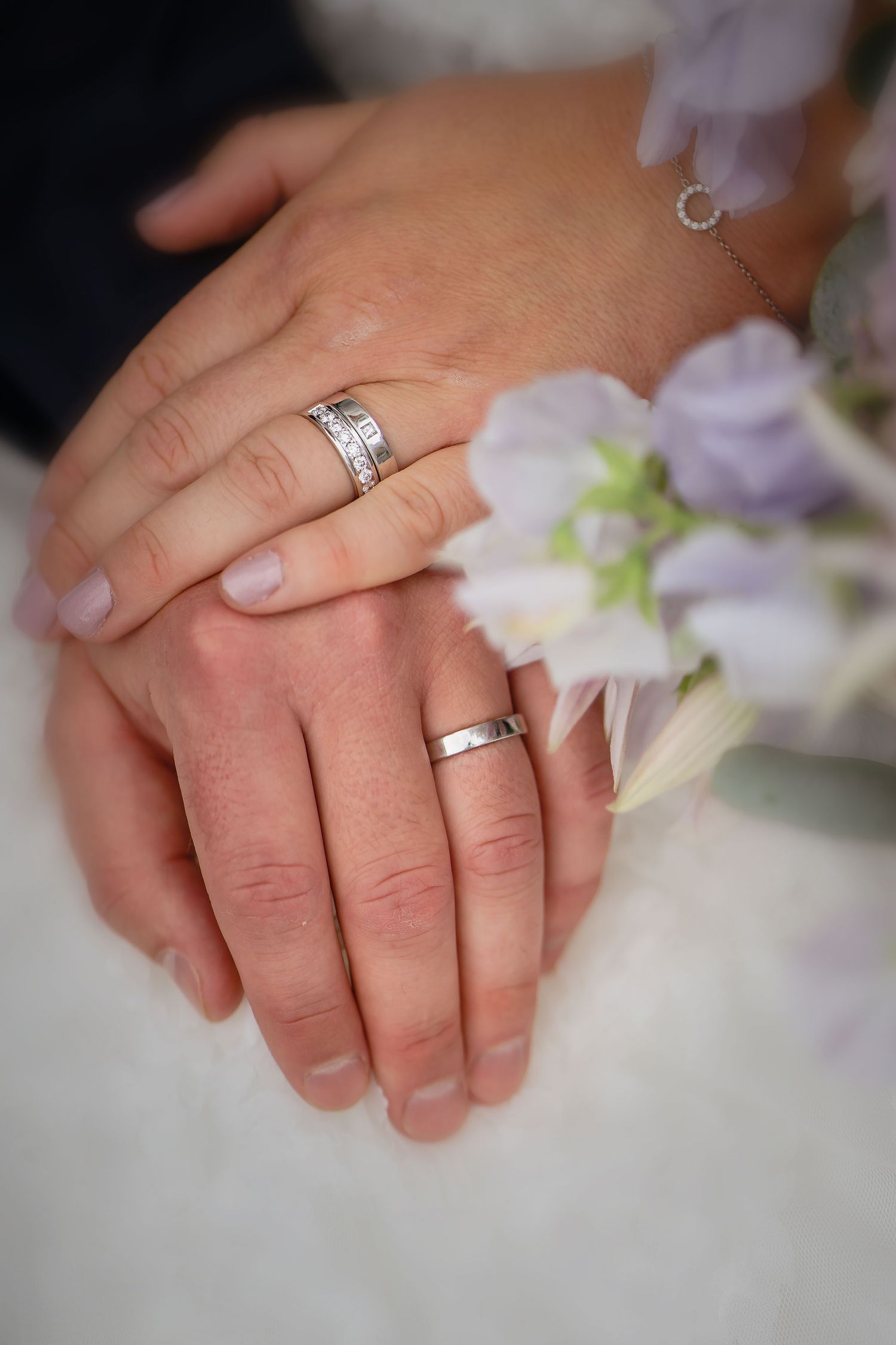 Two newlyweds displaying their silver wedding rings on joined hands beside a soft purple floral bouquet.