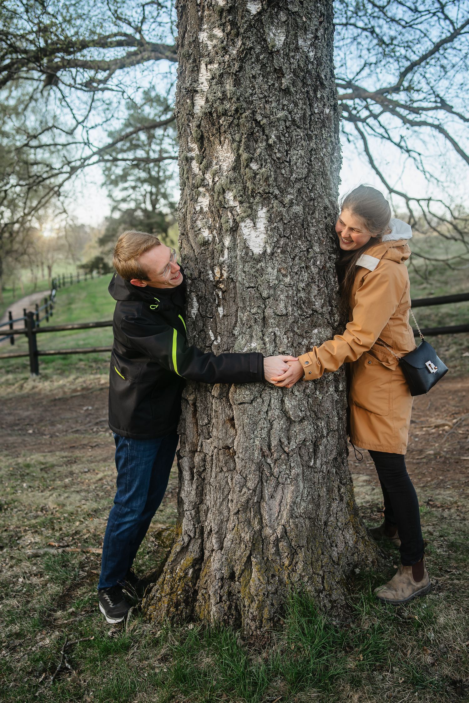 Couple holding hands around a large tree trunk in a park, smiling at each other on a cool autumn day.