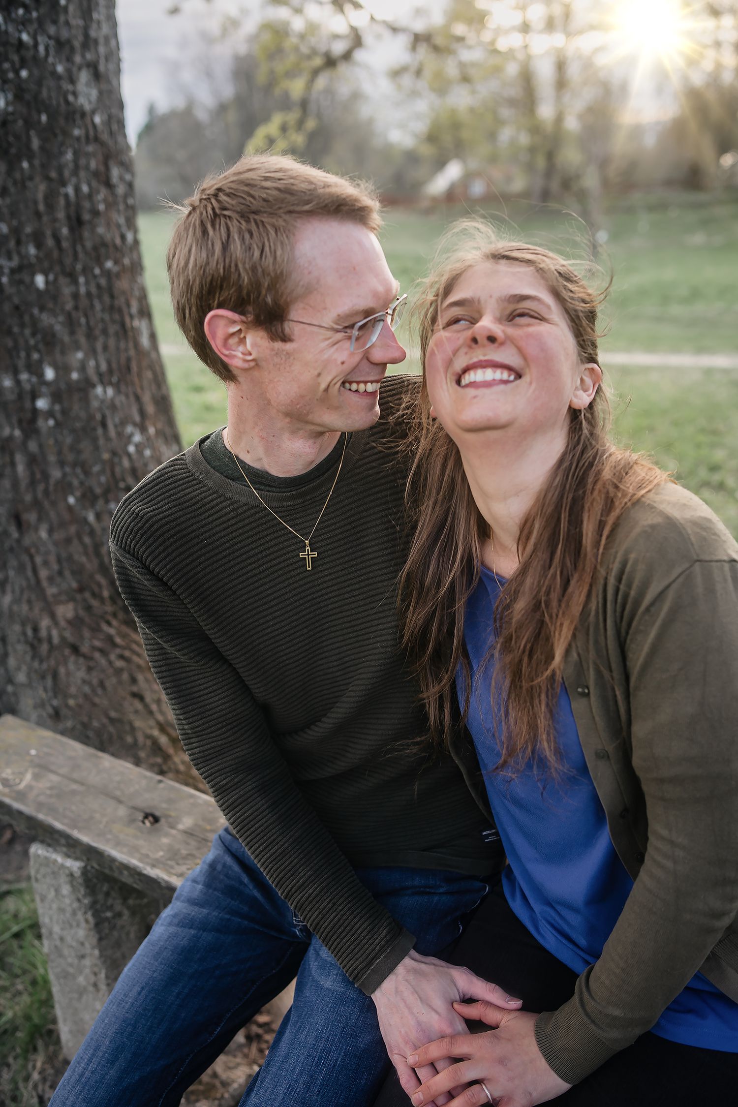 Happy couple laughing together outdoors on a bench, man wearing cross necklace, woman in blue top, golden hour sunlight.