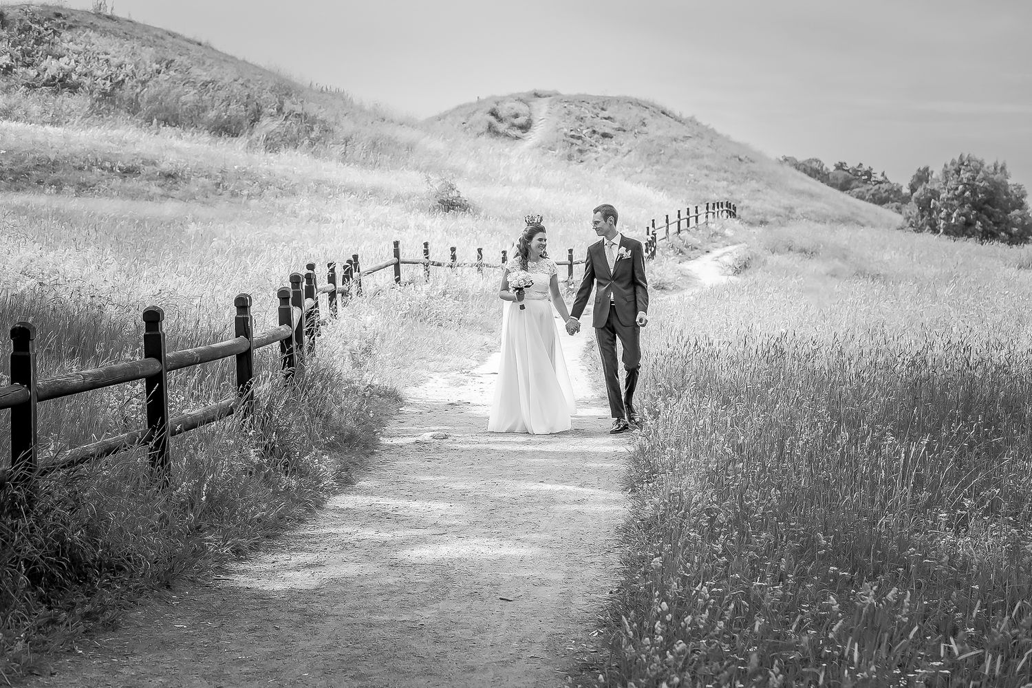 Black and white photo of a bride and groom walking hand in hand along a scenic dirt path with rolling hills.