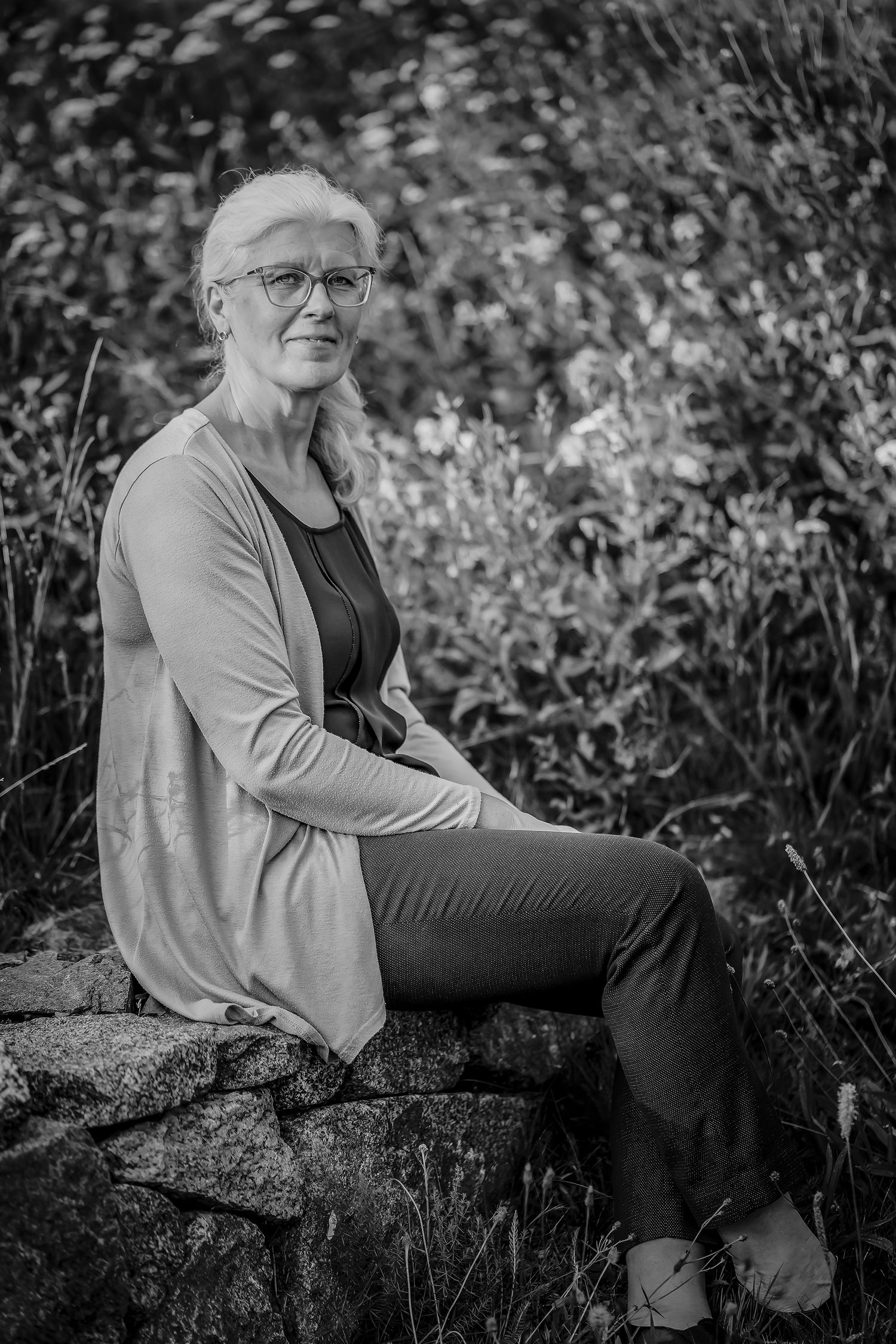 Black and white portrait of an older woman with glasses sitting on a stone wall surrounded by lush garden foliage.