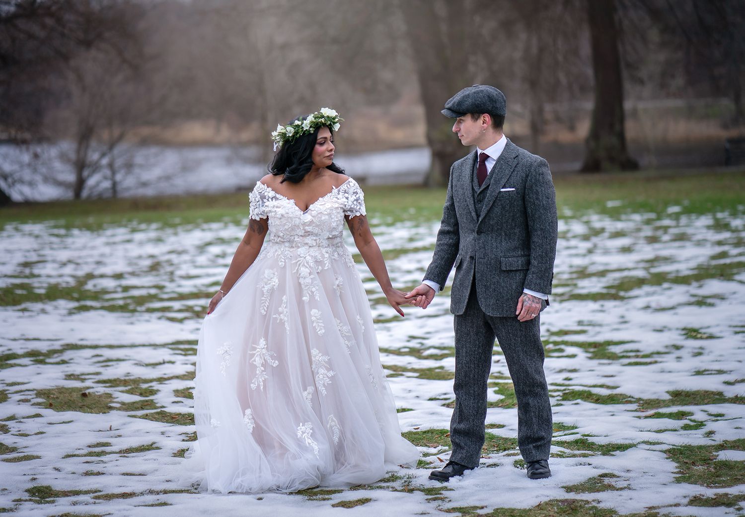 Bride and groom holding hands in snowy park, bride in lace gown with floral crown, groom in grey tweed suit.
