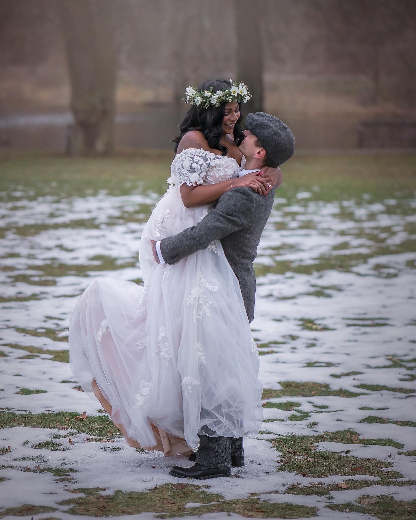 Groom lifts bride in white lace dress and floral crown during romantic winter wedding photo outdoors.