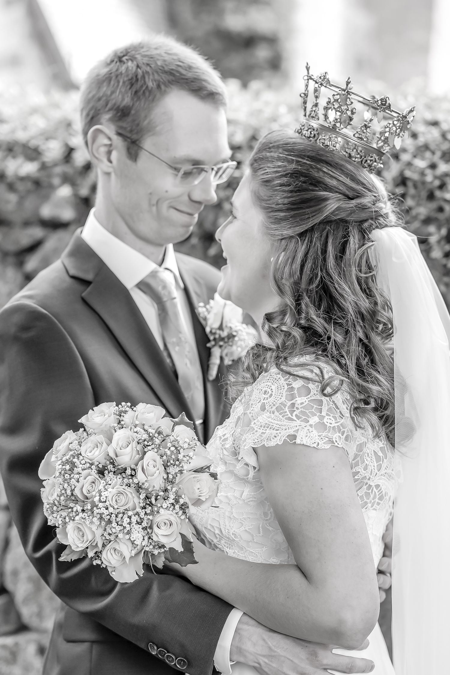 Black and white wedding photo of smiling bride wearing crown holding bouquet with groom in suit and glasses.