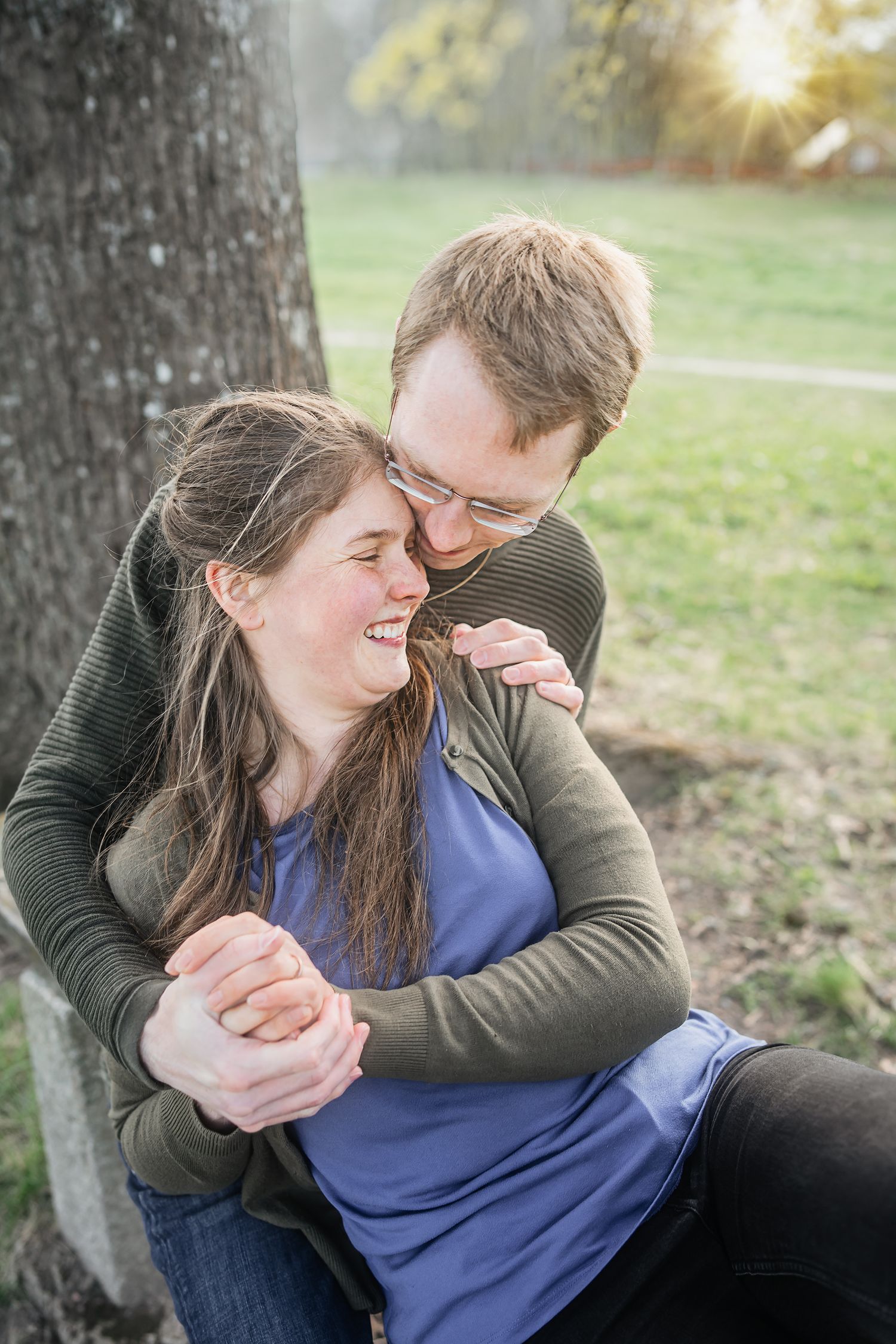 A couple laughs and embraces outdoors near a tree, with warm sunlight glowing behind them in a park setting.
