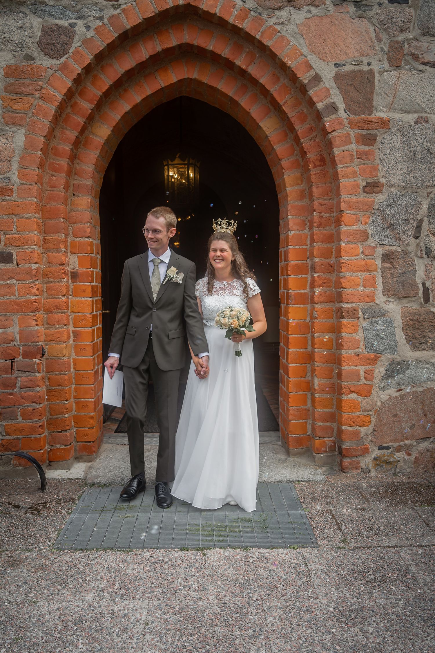 Bride and groom smiling at church doorway, bride wearing crown and white gown holding bouquet, groom in gray suit.