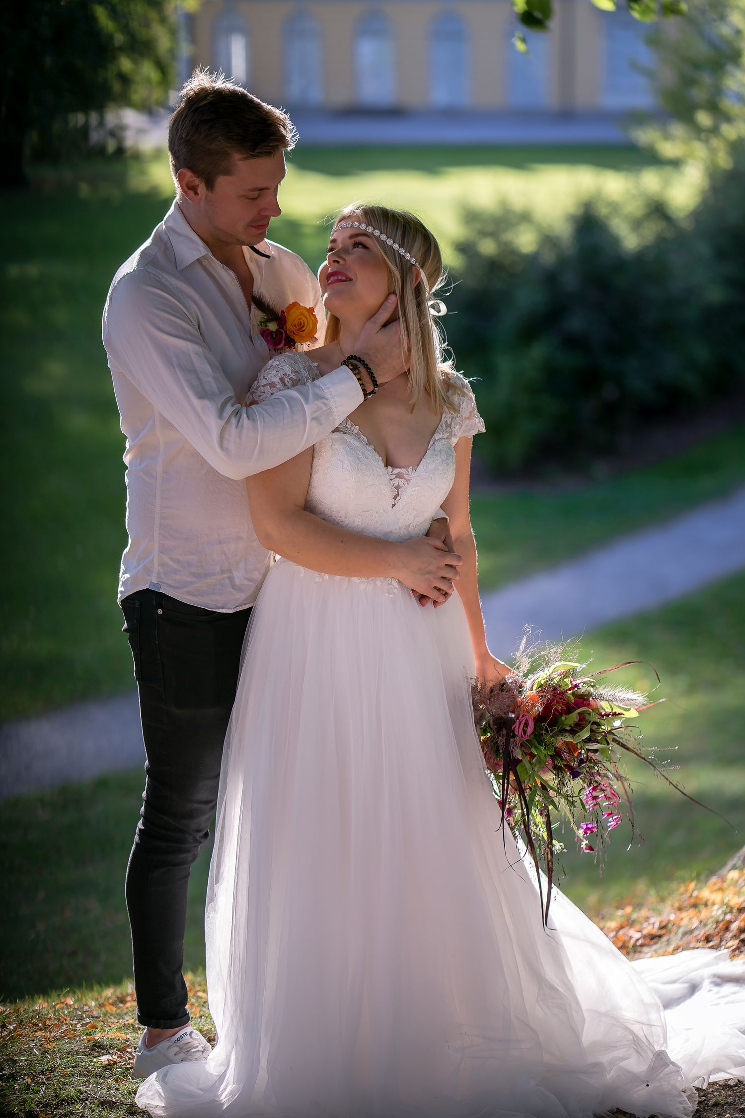A smiling bride in a white tulle gown holding a bouquet, embraced by her groom outdoors in sunlight.