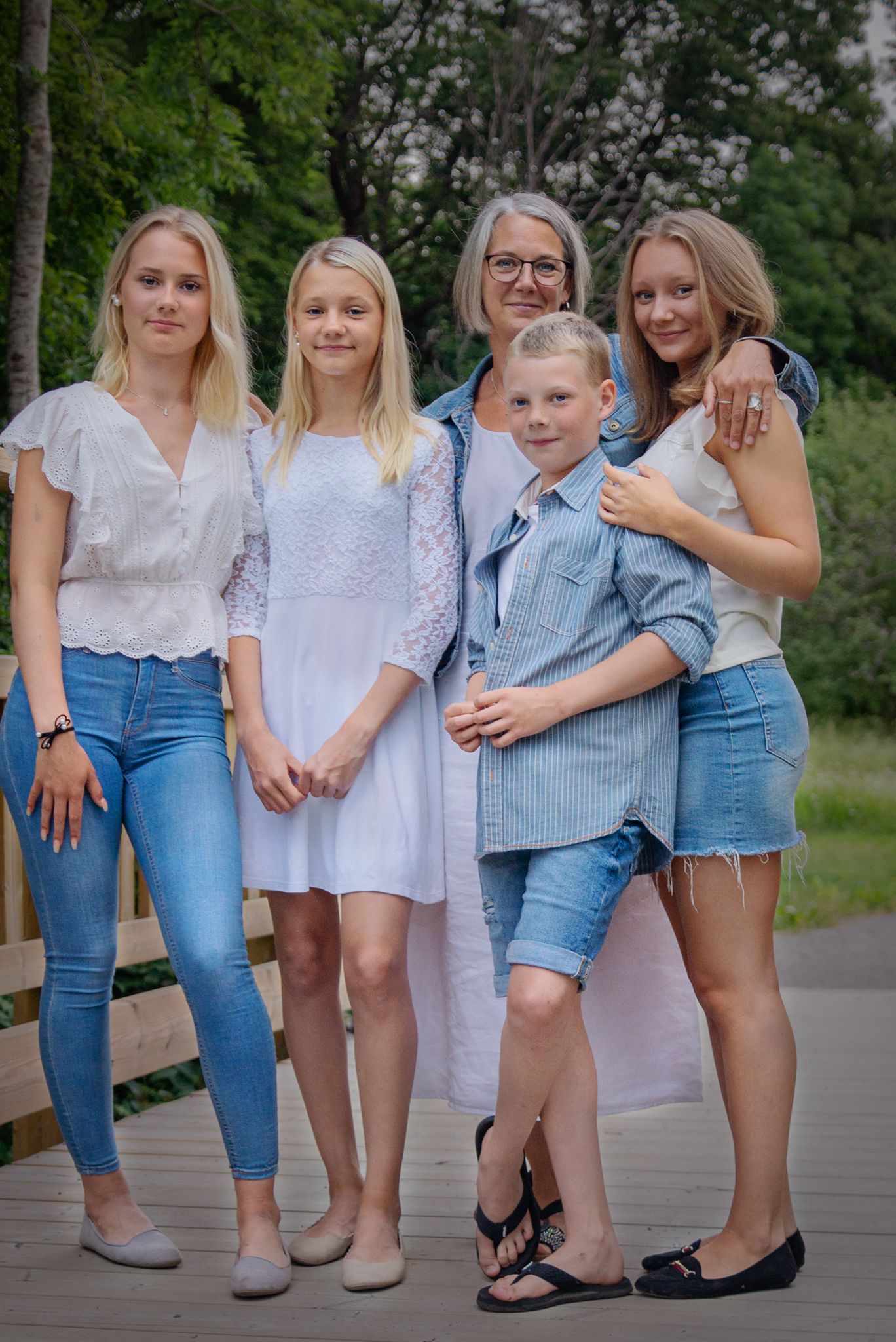 Familjeporträtt av en mamma med fyra barn som poserar på en träbro omgiven av gröna träd.