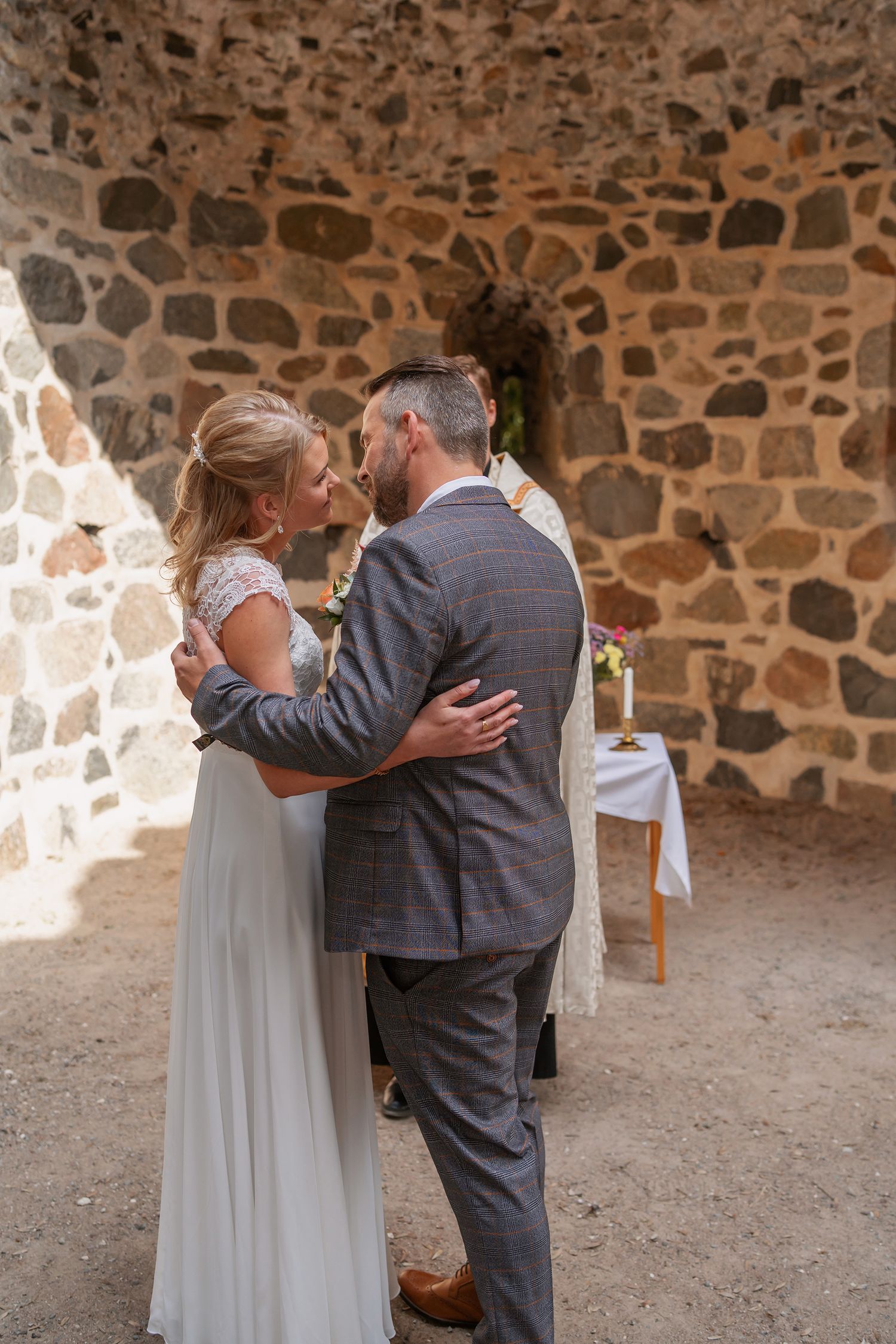 Bride and groom share first kiss during intimate wedding ceremony in rustic stone chapel with officiant behind them.