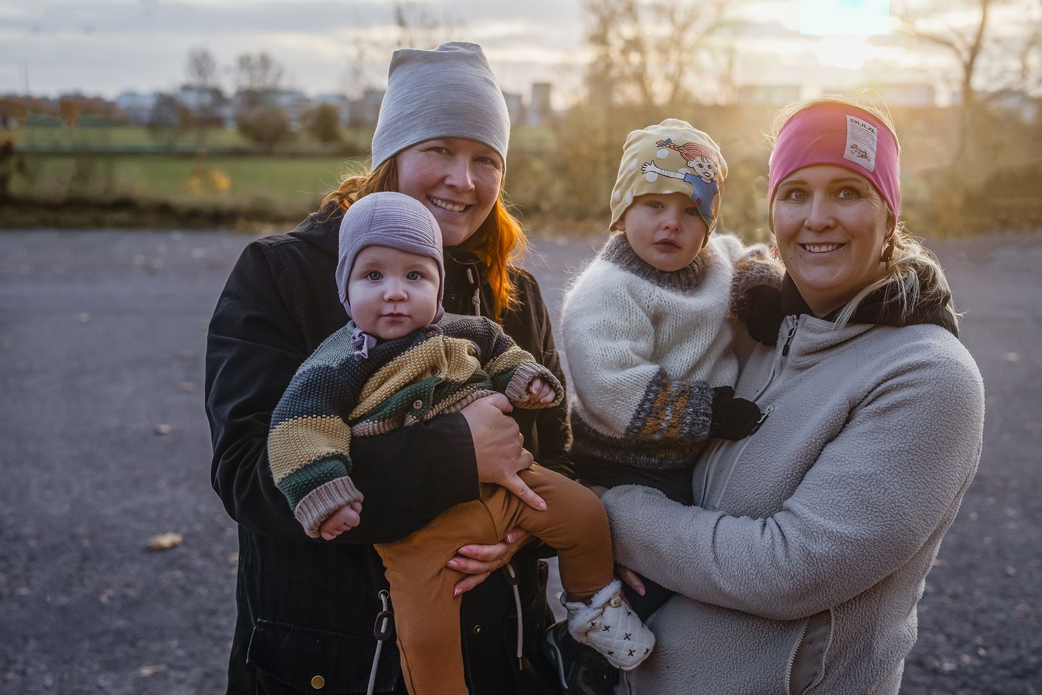 Two women holding babies outdoors at sunset, wearing winter hats, smiling warmly in a rural setting.
