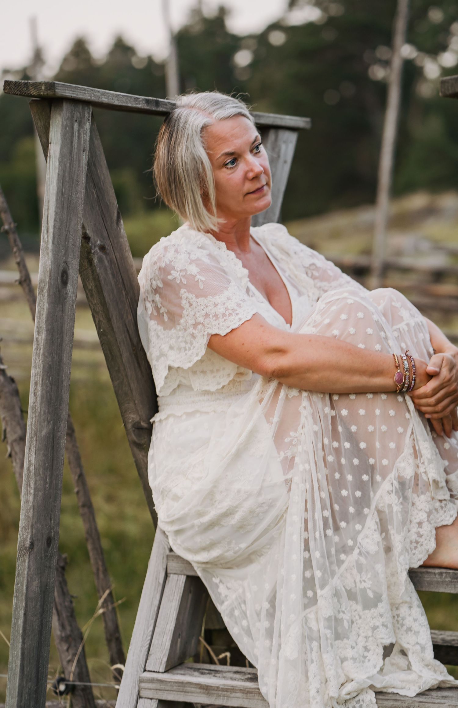 Elegant kvinna i vit spetsklänning som sitter på en trästege i en rustik utomhusmiljö.