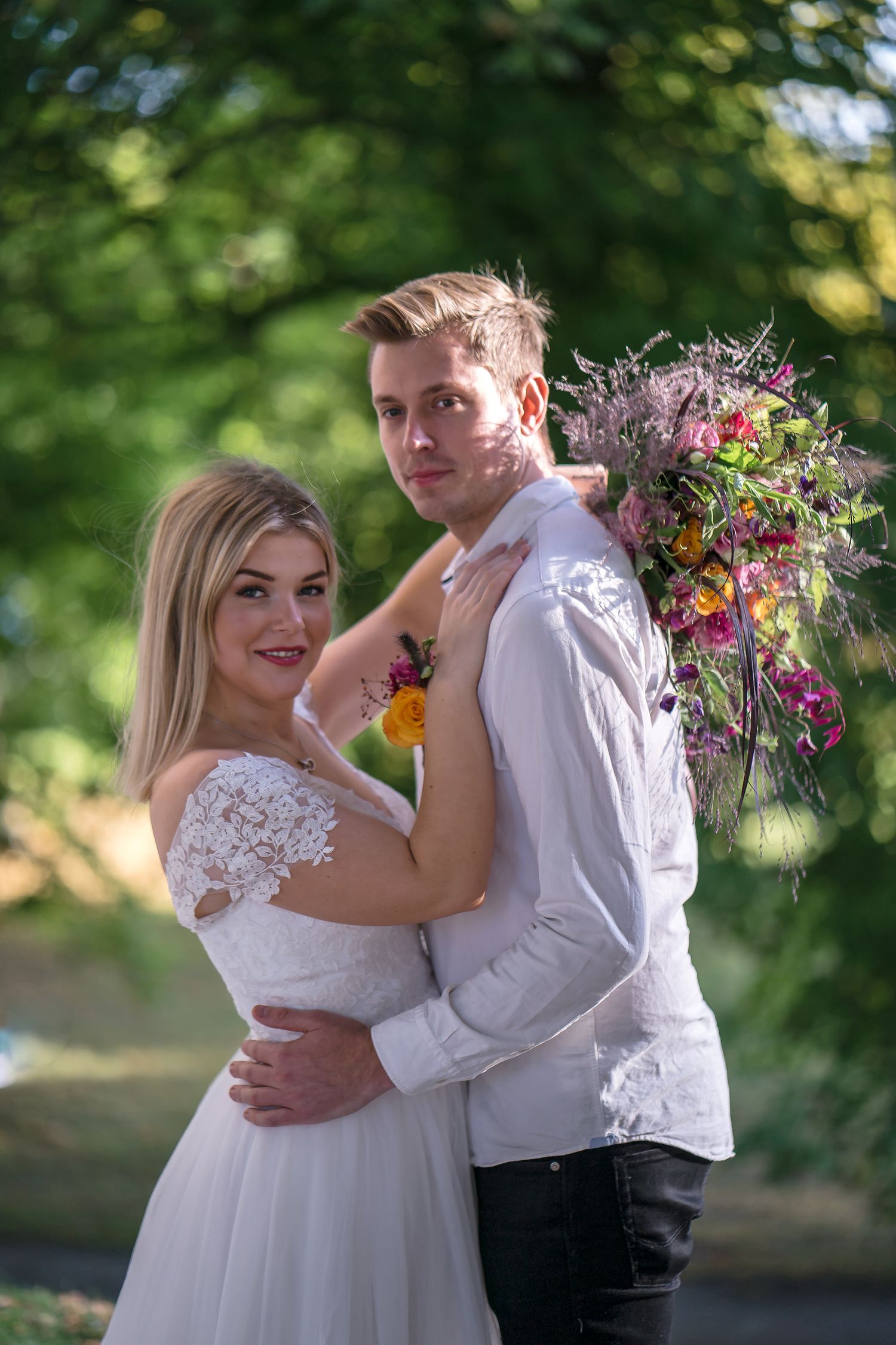 A bride in white lace gown embraces a groom holding a colorful bouquet outdoors with lush green trees.