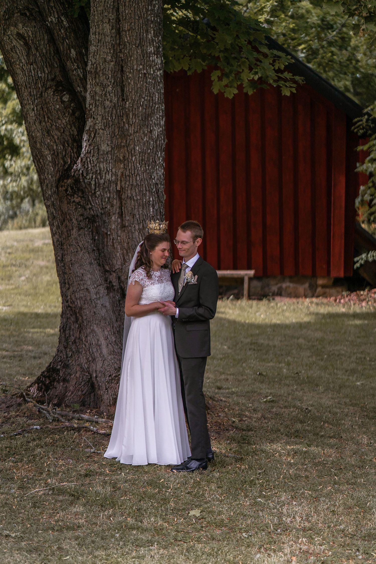 Bride and groom embrace under a large tree with a red barn in the background on their wedding day.