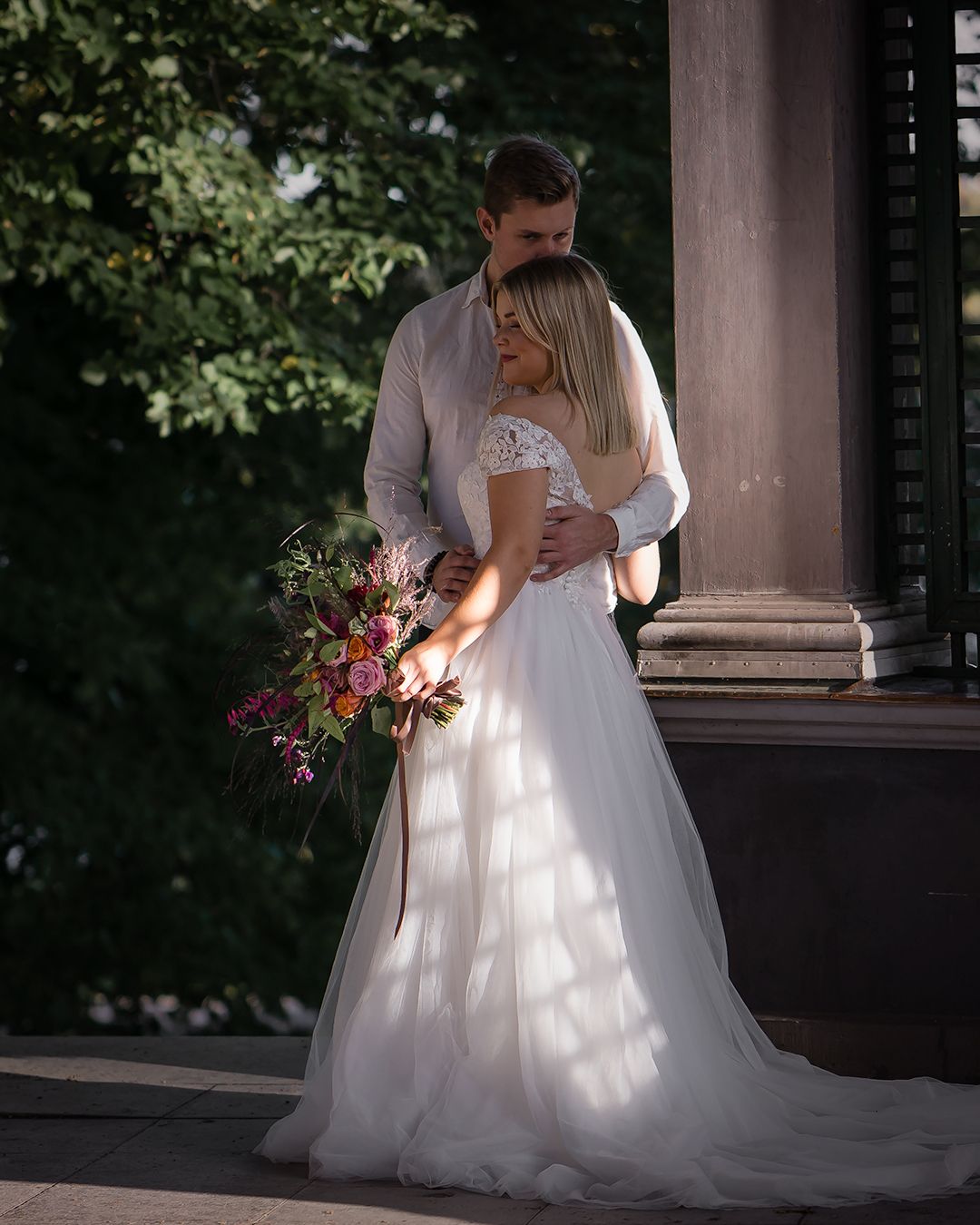 A bride in a white lace gown holds a colorful bouquet while a groom embraces her near stone columns outdoors.
