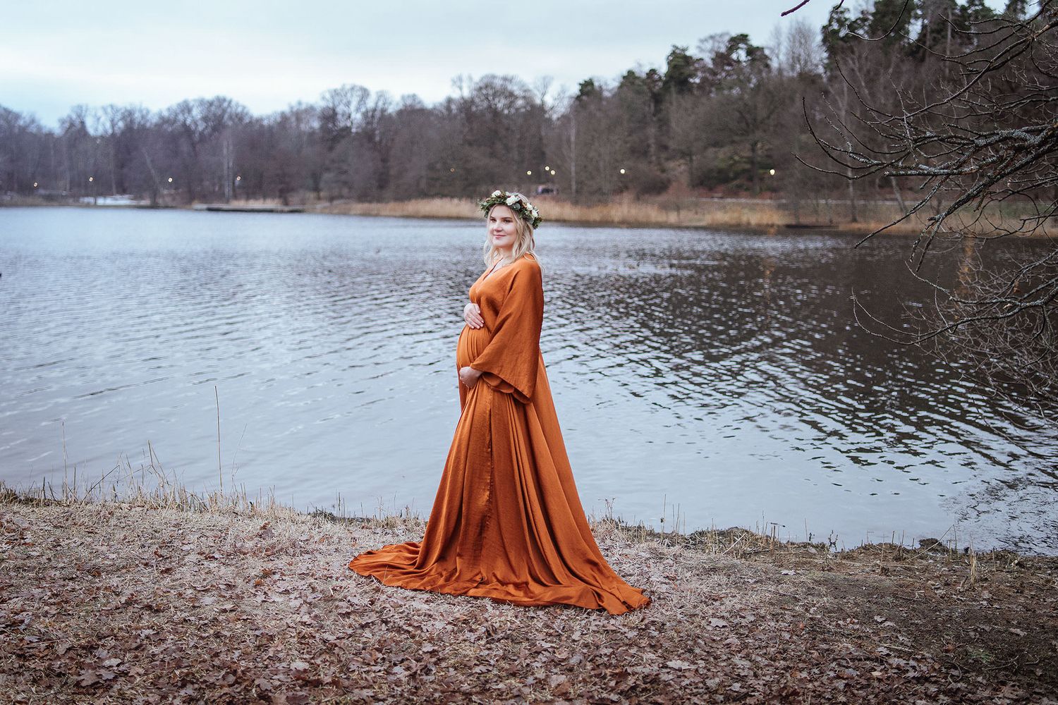 Pregnant woman in flowing rust orange gown with floral crown poses gracefully by a serene lake surrounded by bare trees.