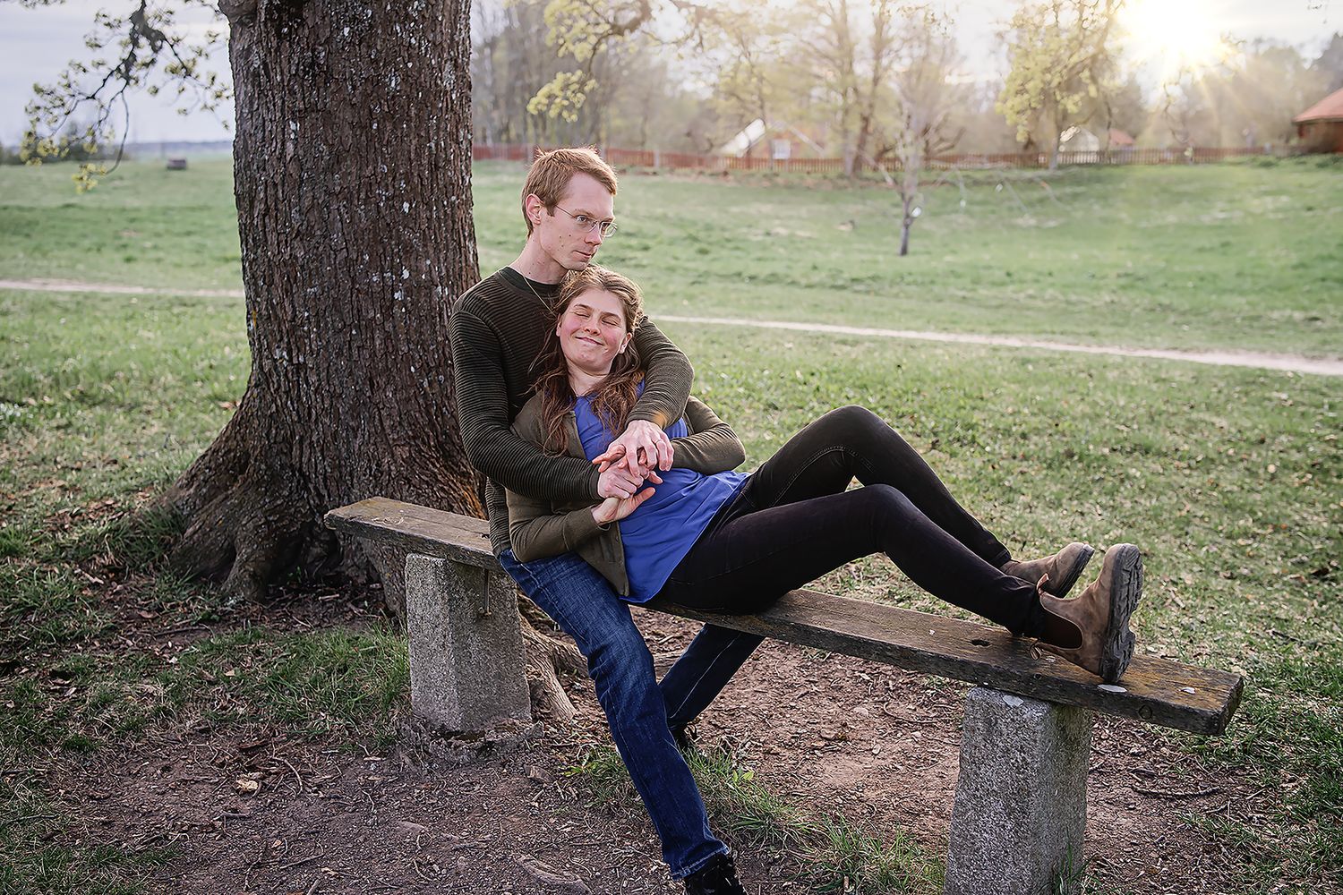 Couple relaxing on a wooden park bench under a tree, woman lying across man's lap in a green outdoor setting.