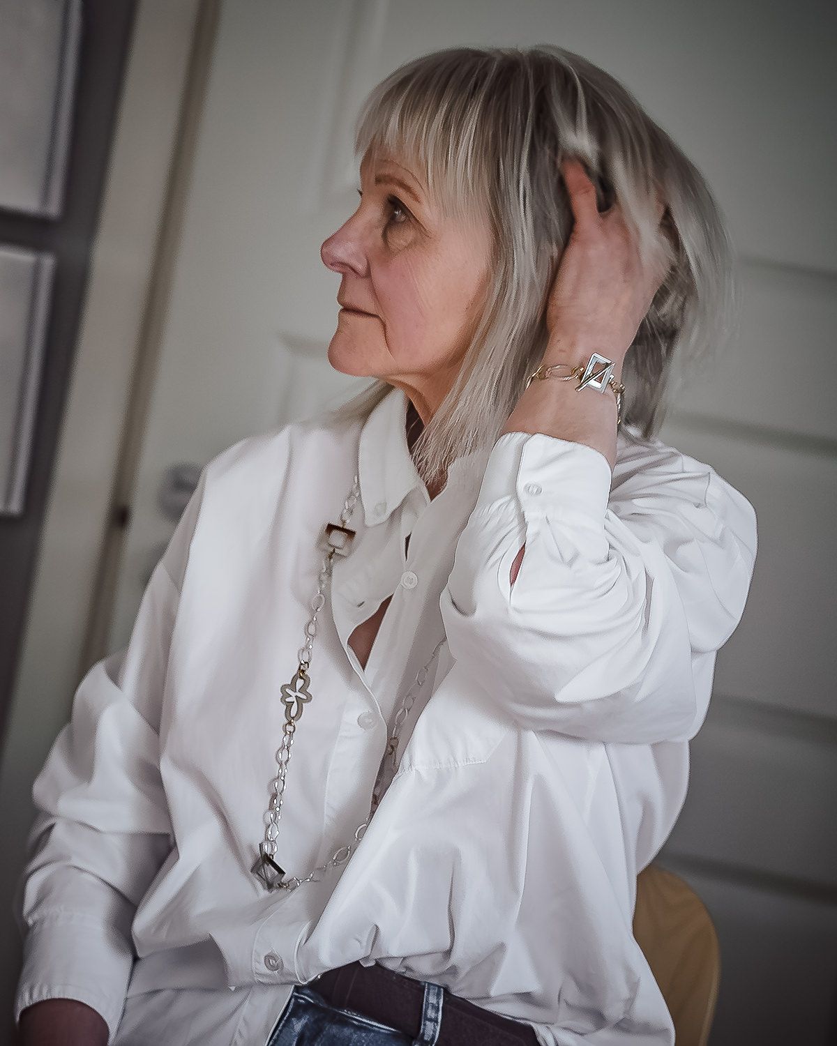 Side profile portrait showing a person with silver hair wearing a crisp white button-up shirt and necklace in soft lighting.