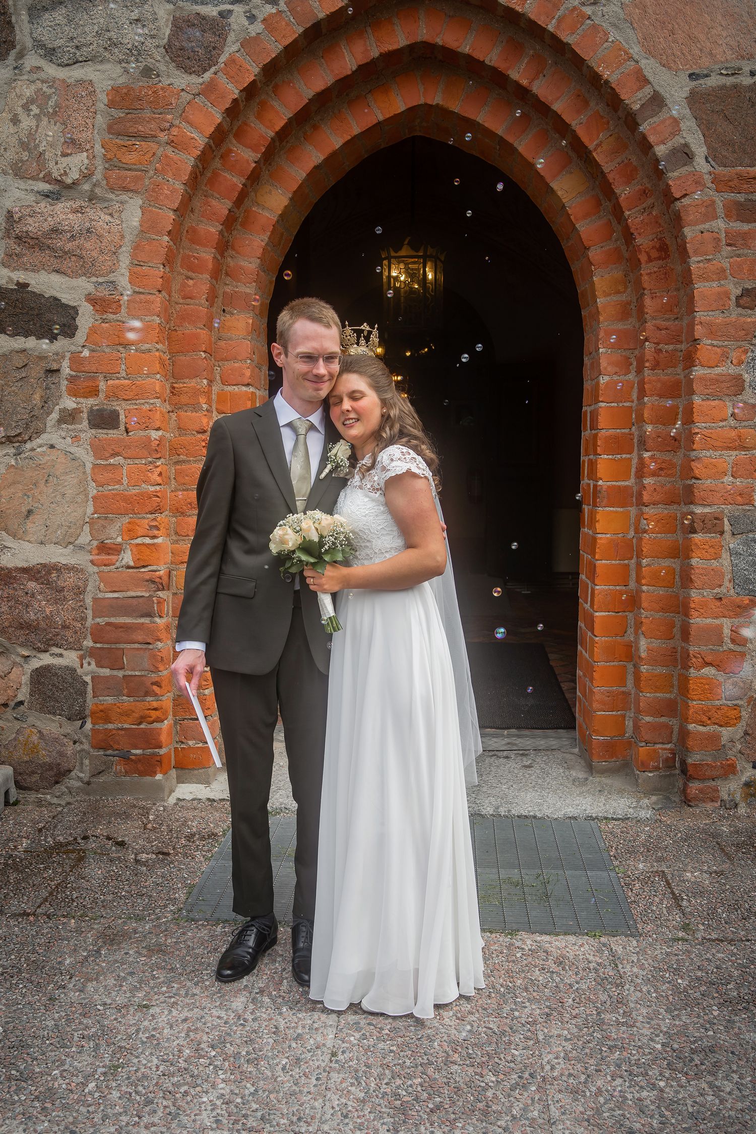 Newlywed couple posing in a brick church archway, bride in white gown holding bouquet, groom in gray suit.