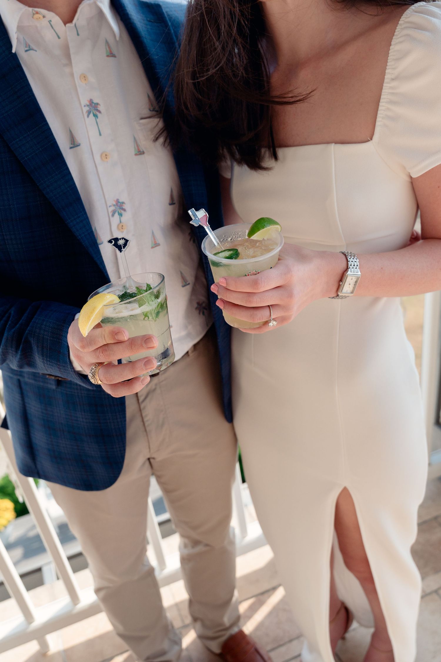 Couple holding mojito cocktails with lime and straws, dressed in elegant wedding attire at an outdoor venue.