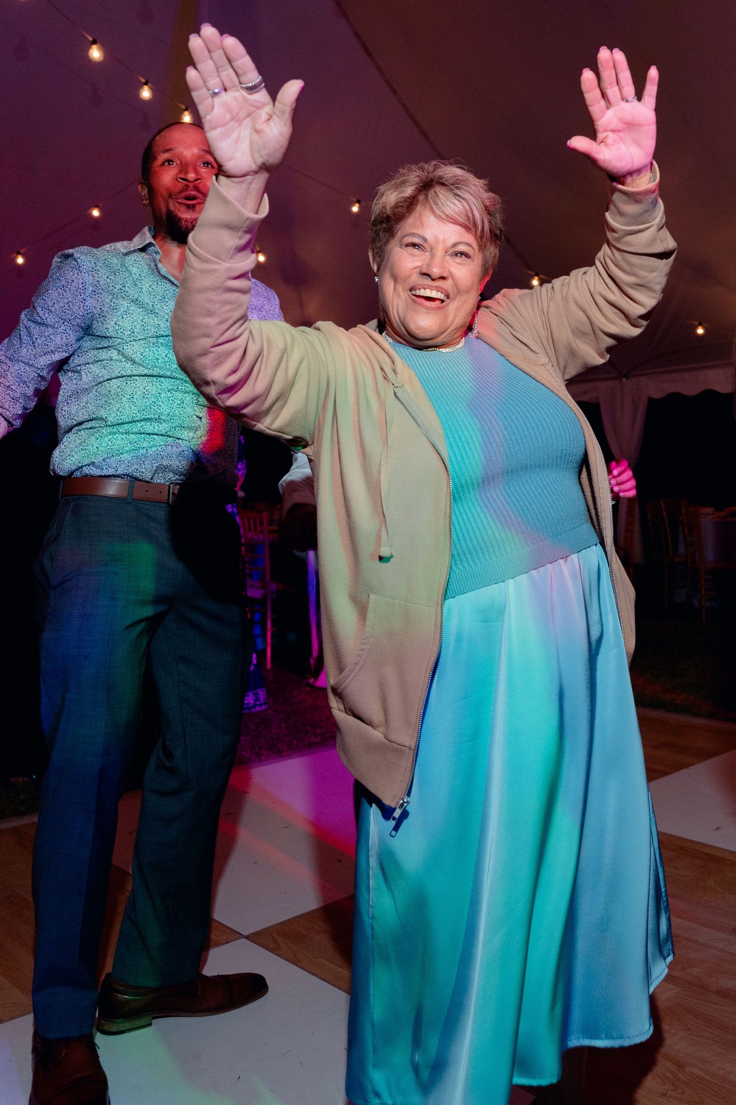 Joyful older woman with hands raised dancing at wedding reception under colorful lights with guests.