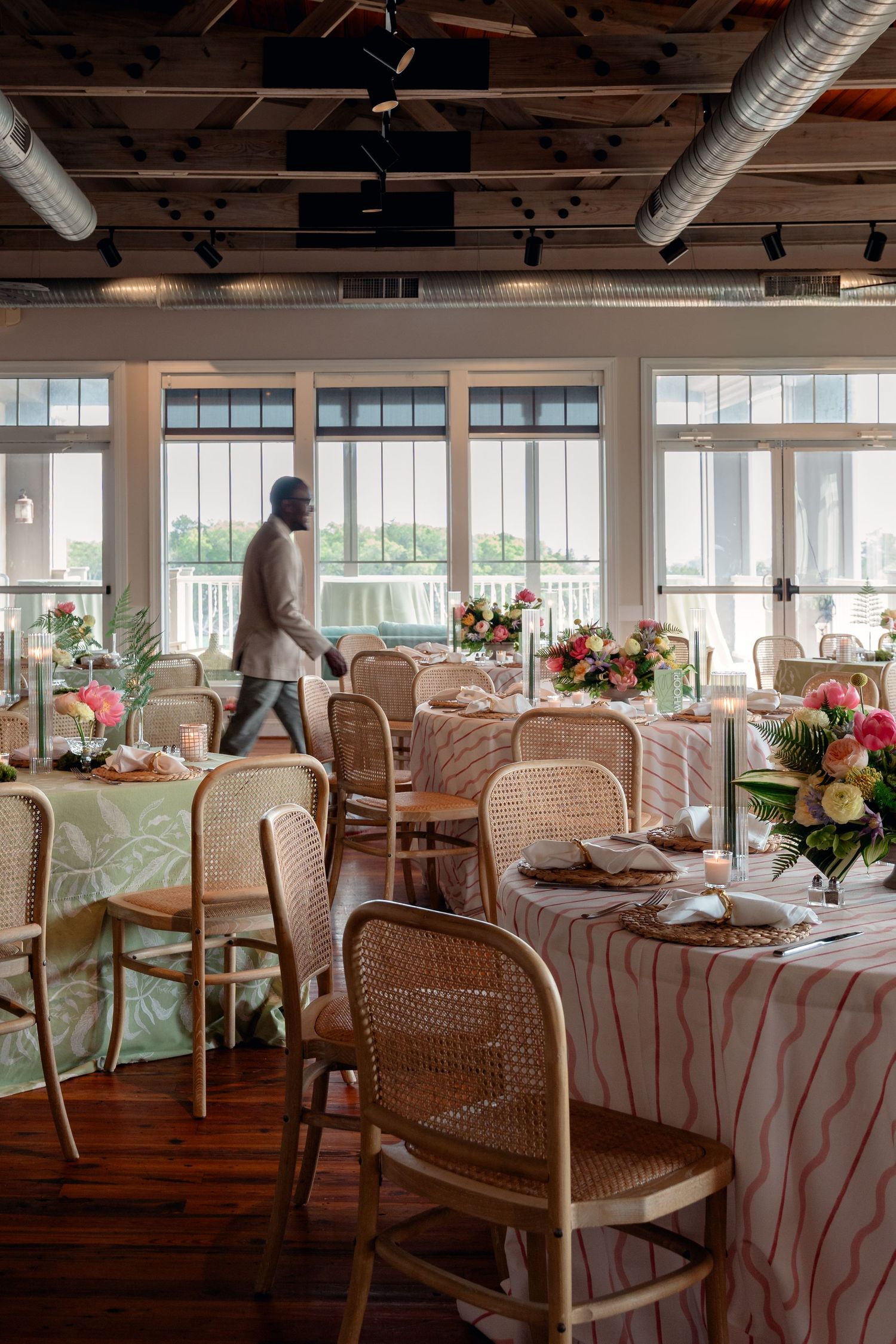 Elegant event venue with rattan chairs, floral centerpieces, and striped tablecloths beside large windows.