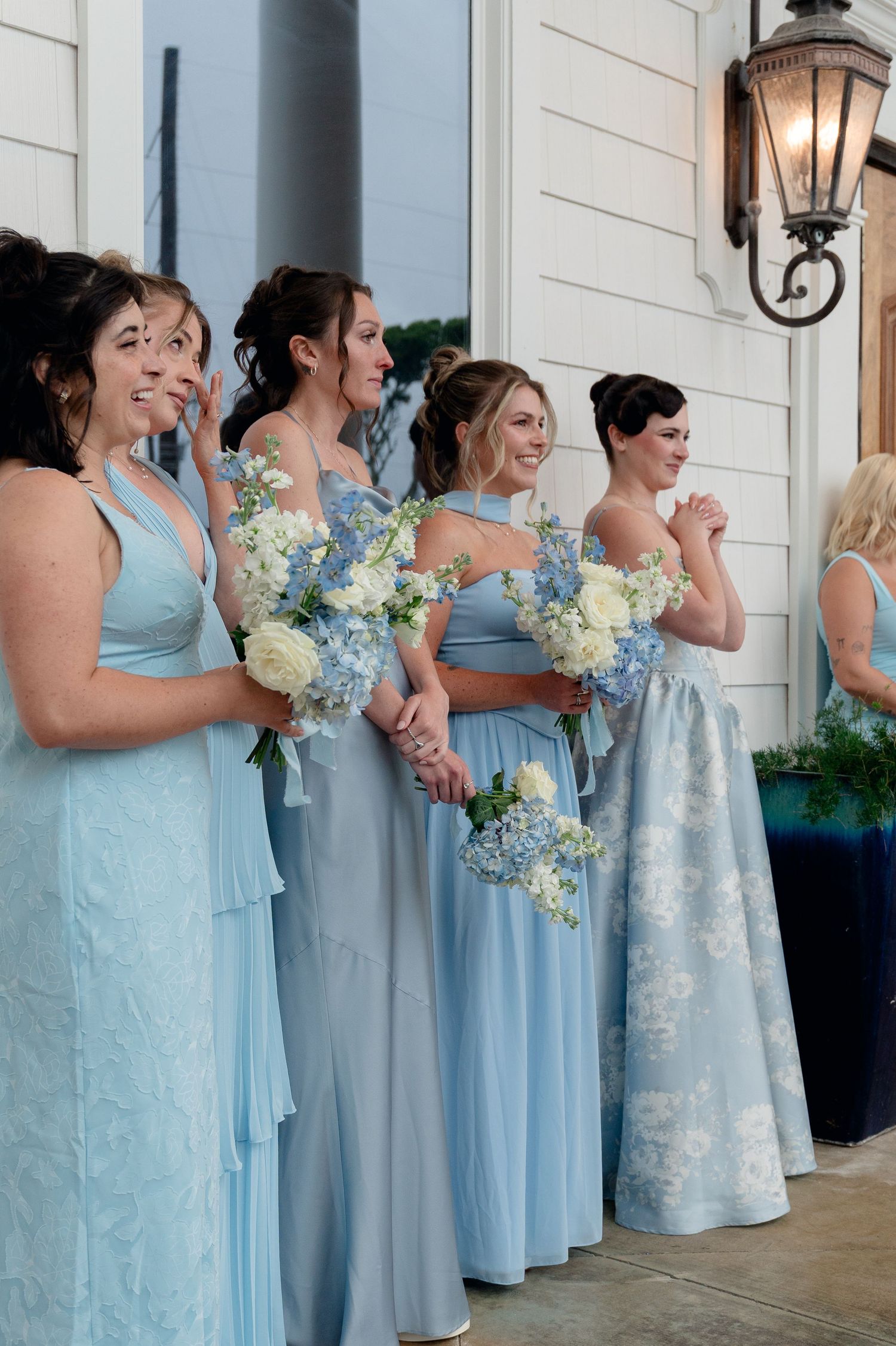 Bridesmaids in light blue gowns smile and laugh while holding white and blue floral bouquets outside white venue entrance.