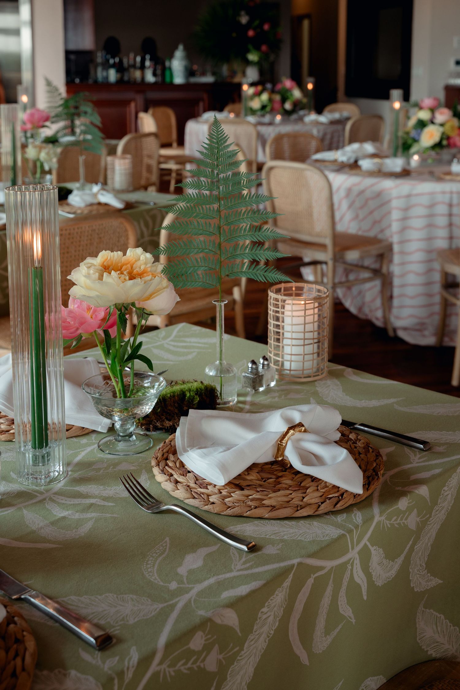 Elegant wedding table setting with wicker charger, white napkin, fern decor, candles, and floral centerpiece on sage tablecloth.