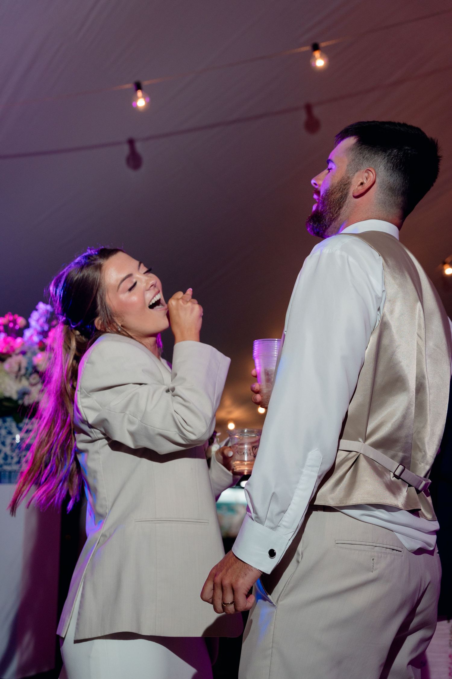 Bride in white blazer and groom in vest laughing and dancing together at wedding reception under string lights.