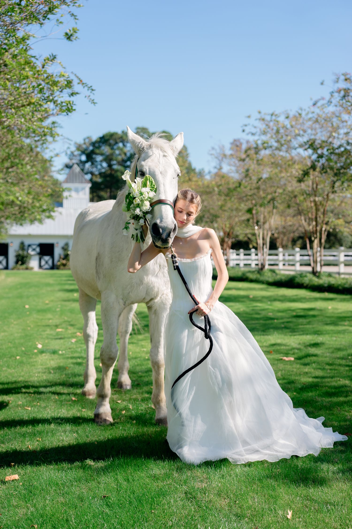 Bride in a strapless white gown posing with a white horse adorned with floral wreath on green lawn.
