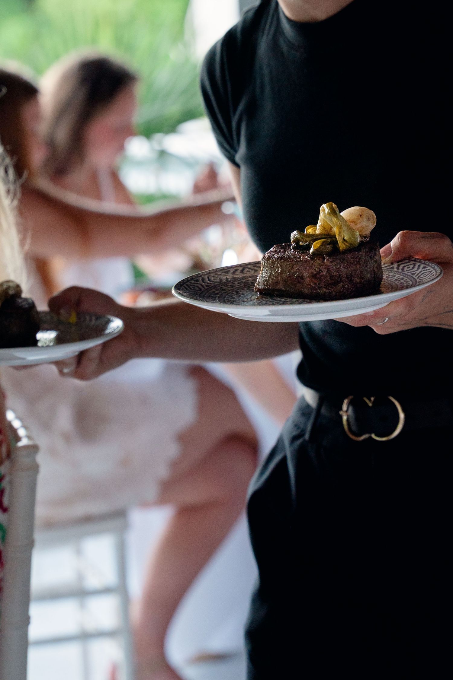 Server in black carrying elegant plate of steak with garnish at upscale catered event.