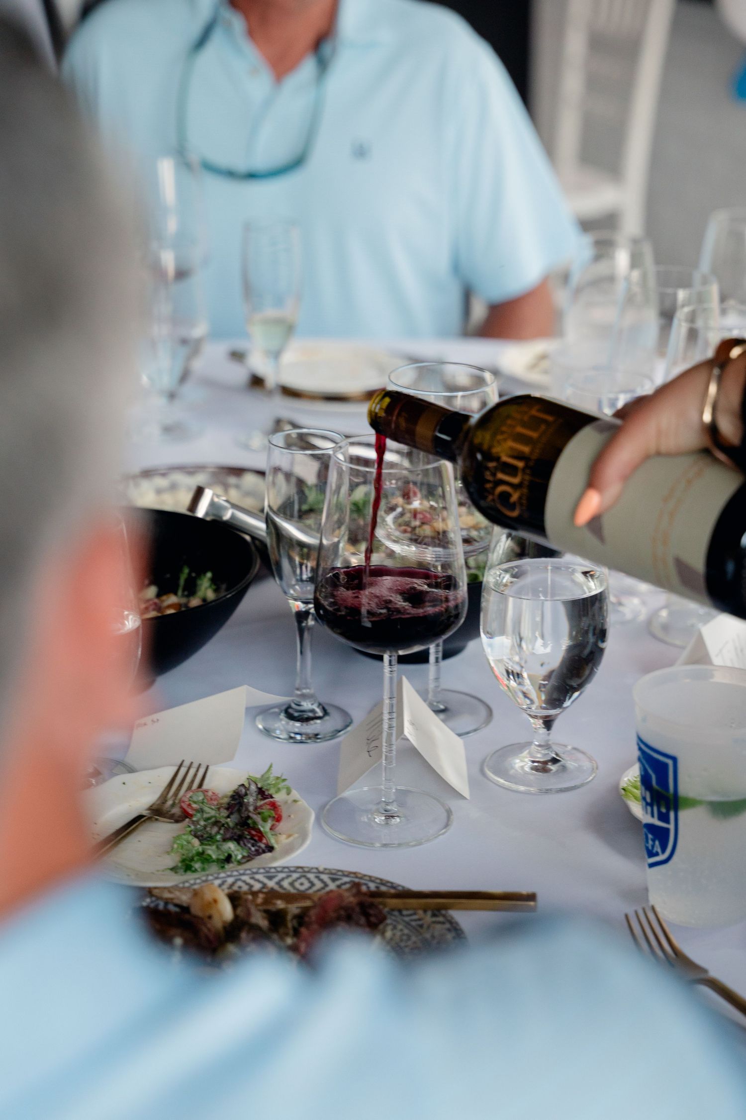 Red wine being poured into a glass at an elegant dinner table setting with white tablecloth and crystal glassware.