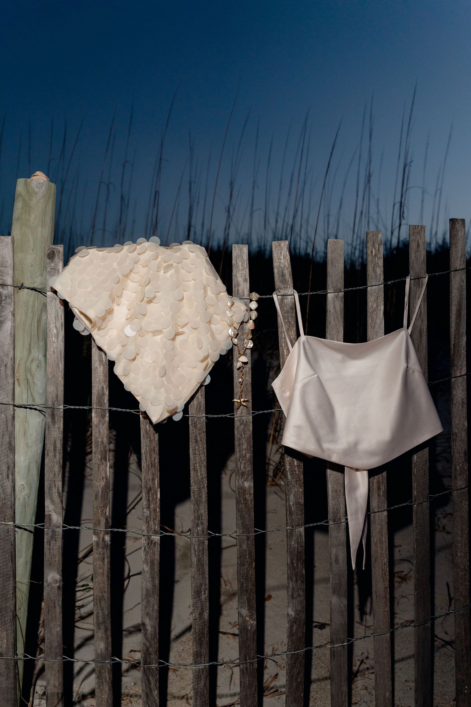 White lace garment and silk camisole hanging on a rustic wooden beach fence at night.
