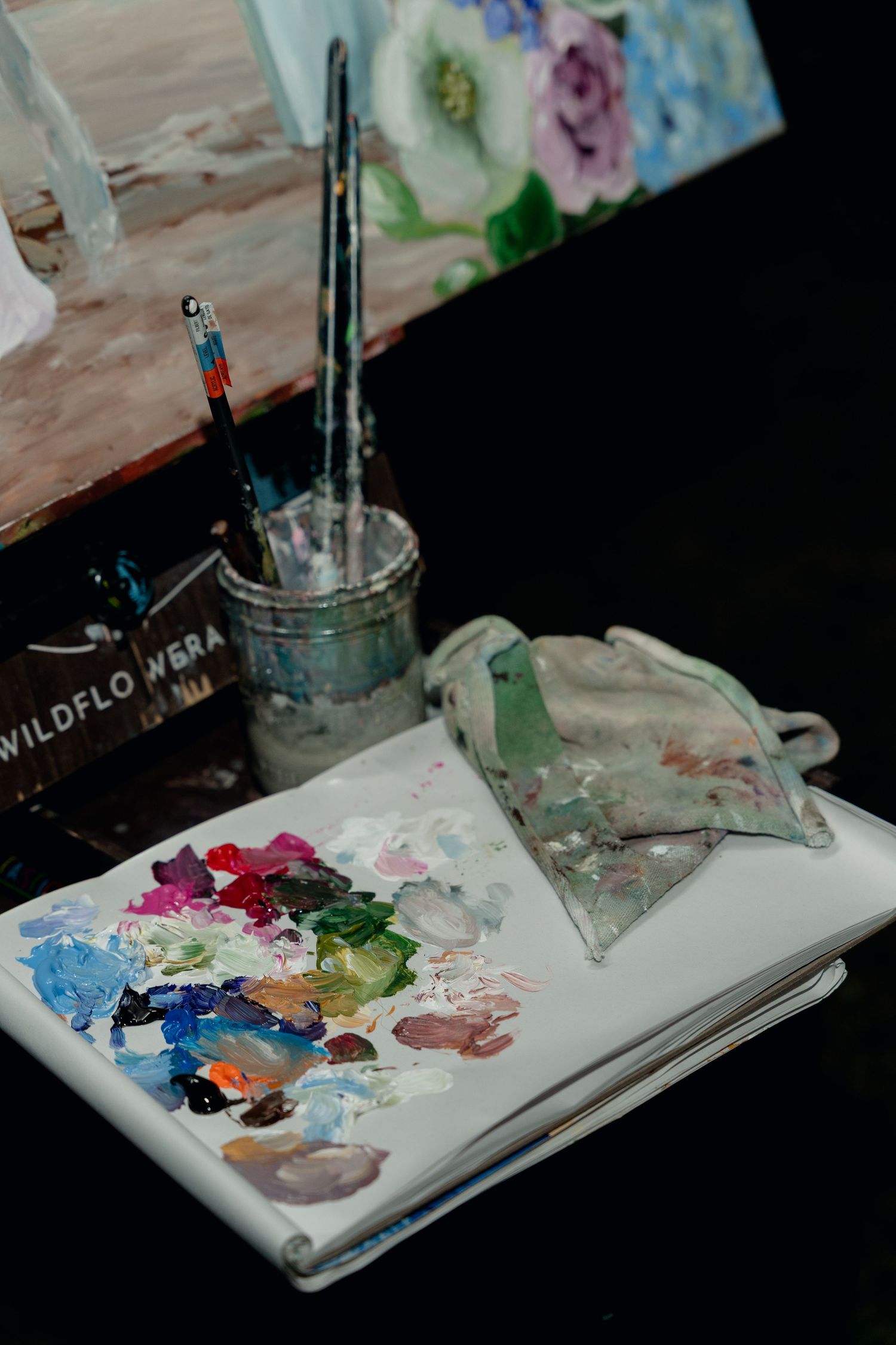 Artist palette and paint brushes on sketchbook beside colorful floral canvas painting at wedding event.