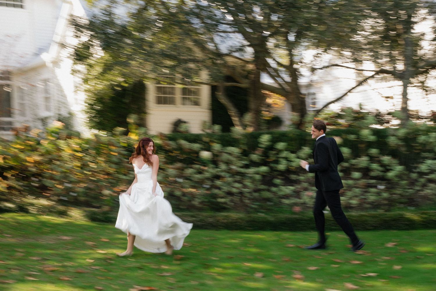 Bride and groom running joyfully across a green lawn outside a white house on their wedding day.