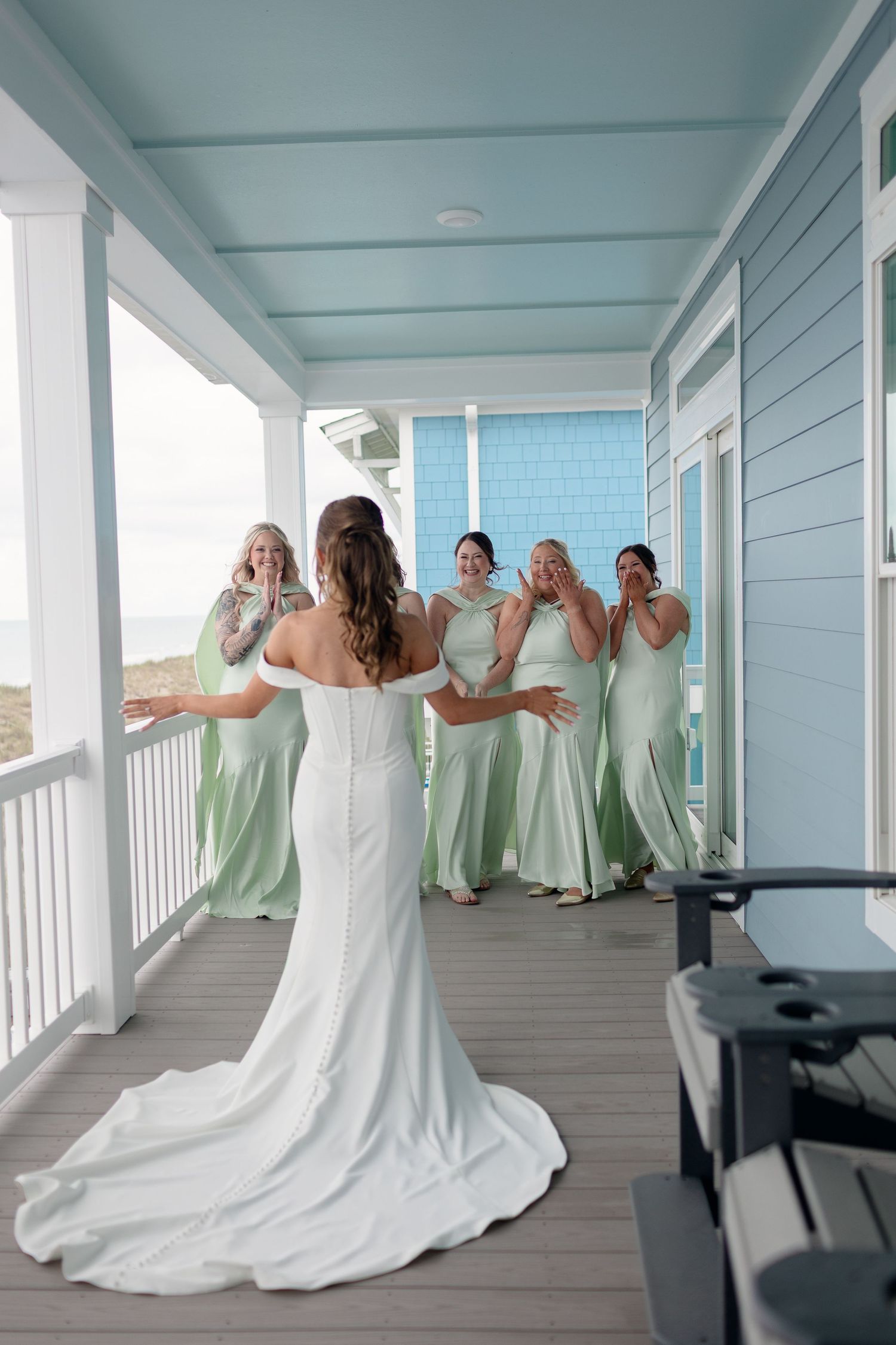 Bride in white gown surprises bridesmaids in mint green dresses on a coastal beach house porch.