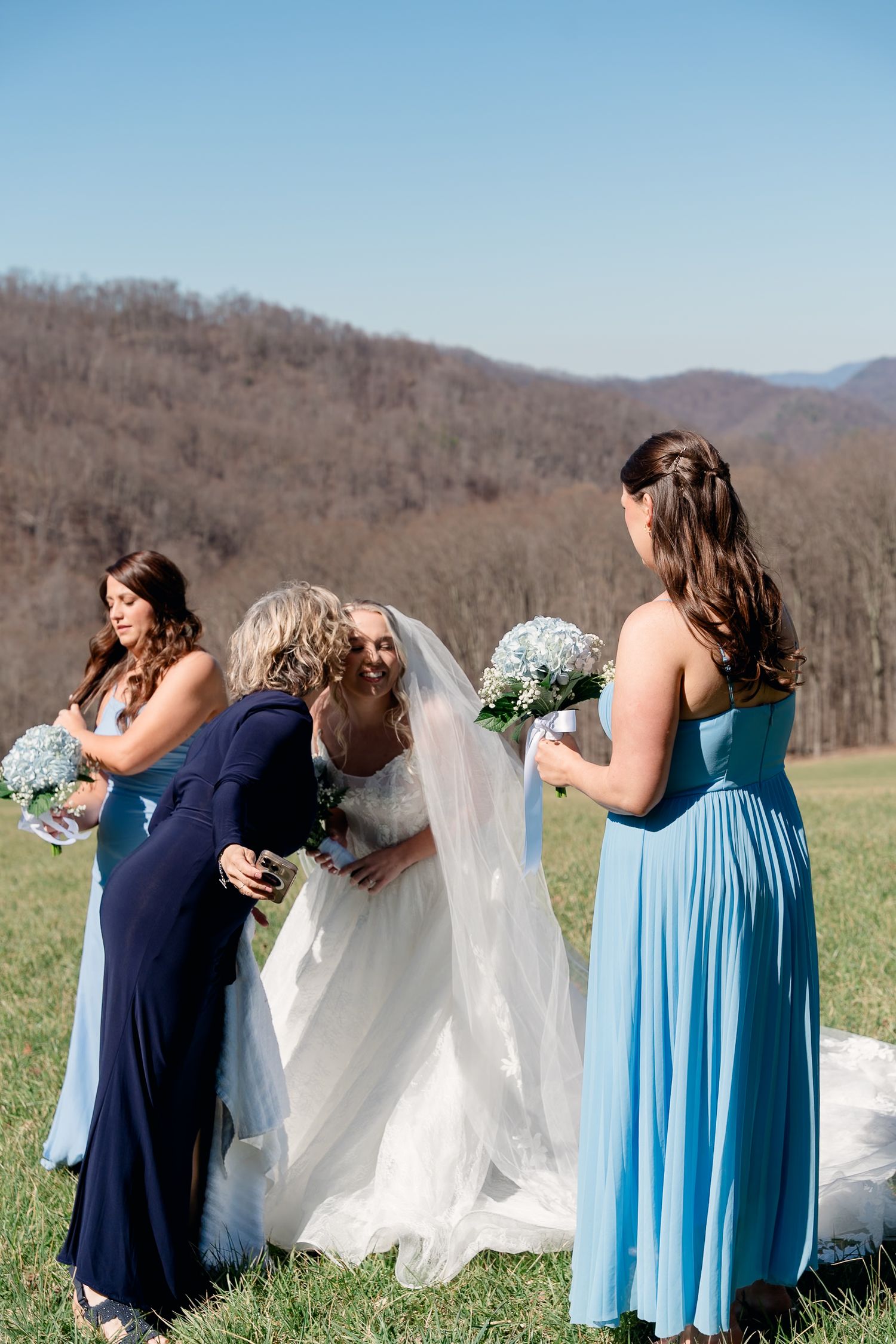 Older woman hugs blonde bride in white gown while bridesmaids in blue dresses look on in scenic mountain setting.