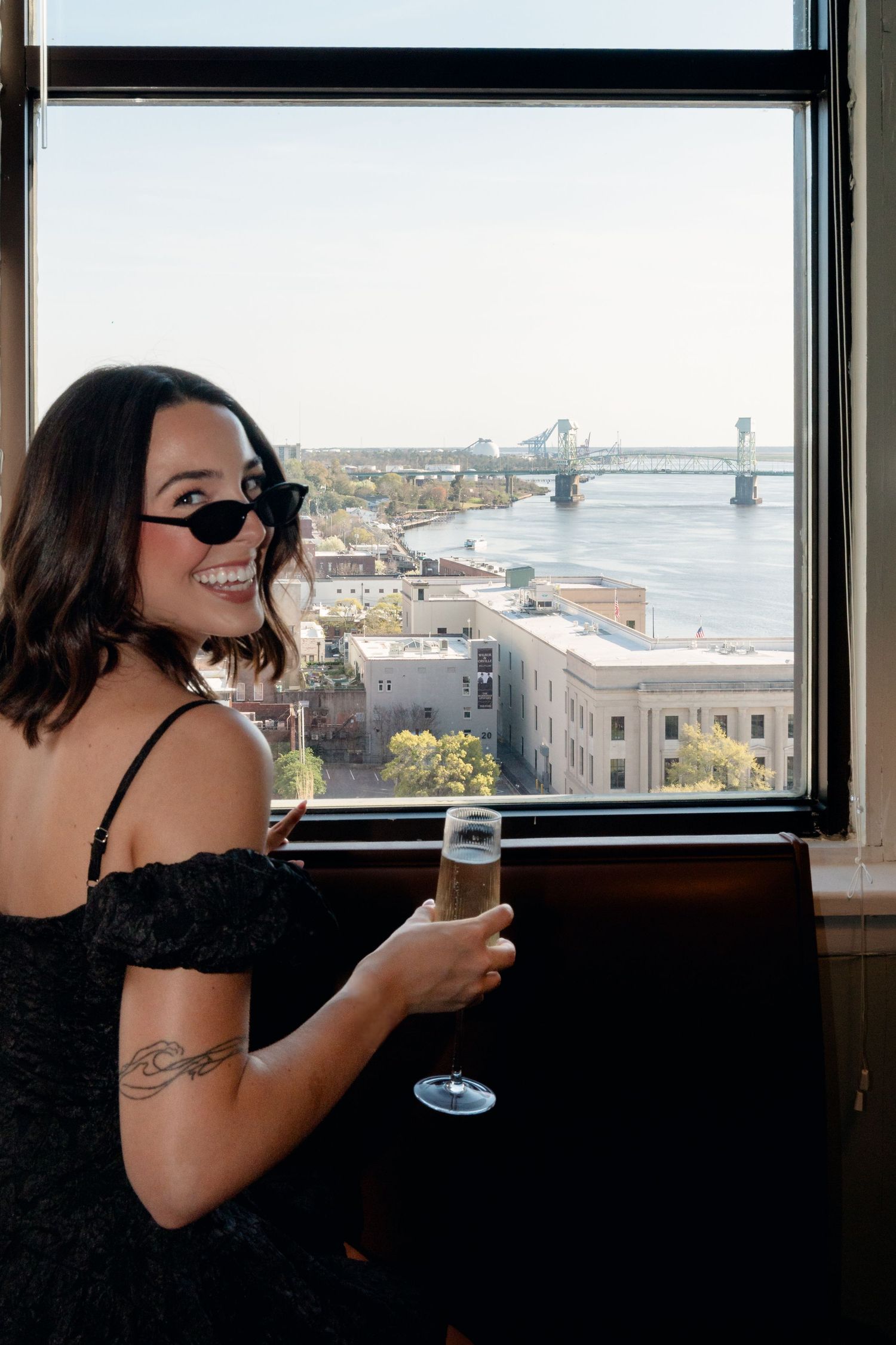 Smiling woman in black dress with sunglasses holding champagne glass by window with city river view.
