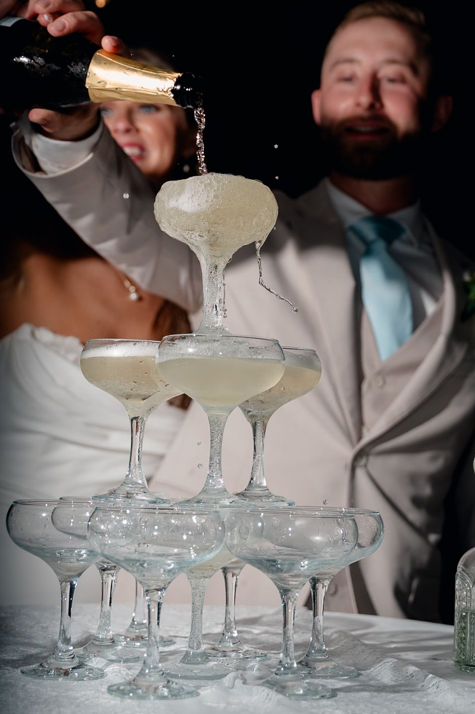 Champagne tower being filled at a wedding reception with bride and groom smiling in the background.