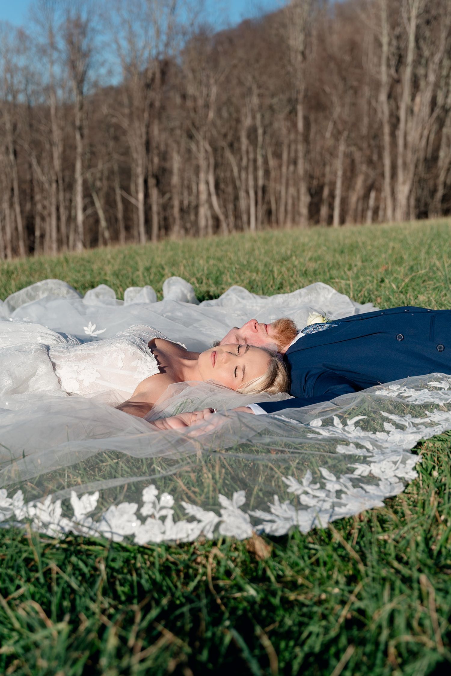 Bride and groom lay together on green grass, bride's white lace veil spread around them with wooded hills behind.